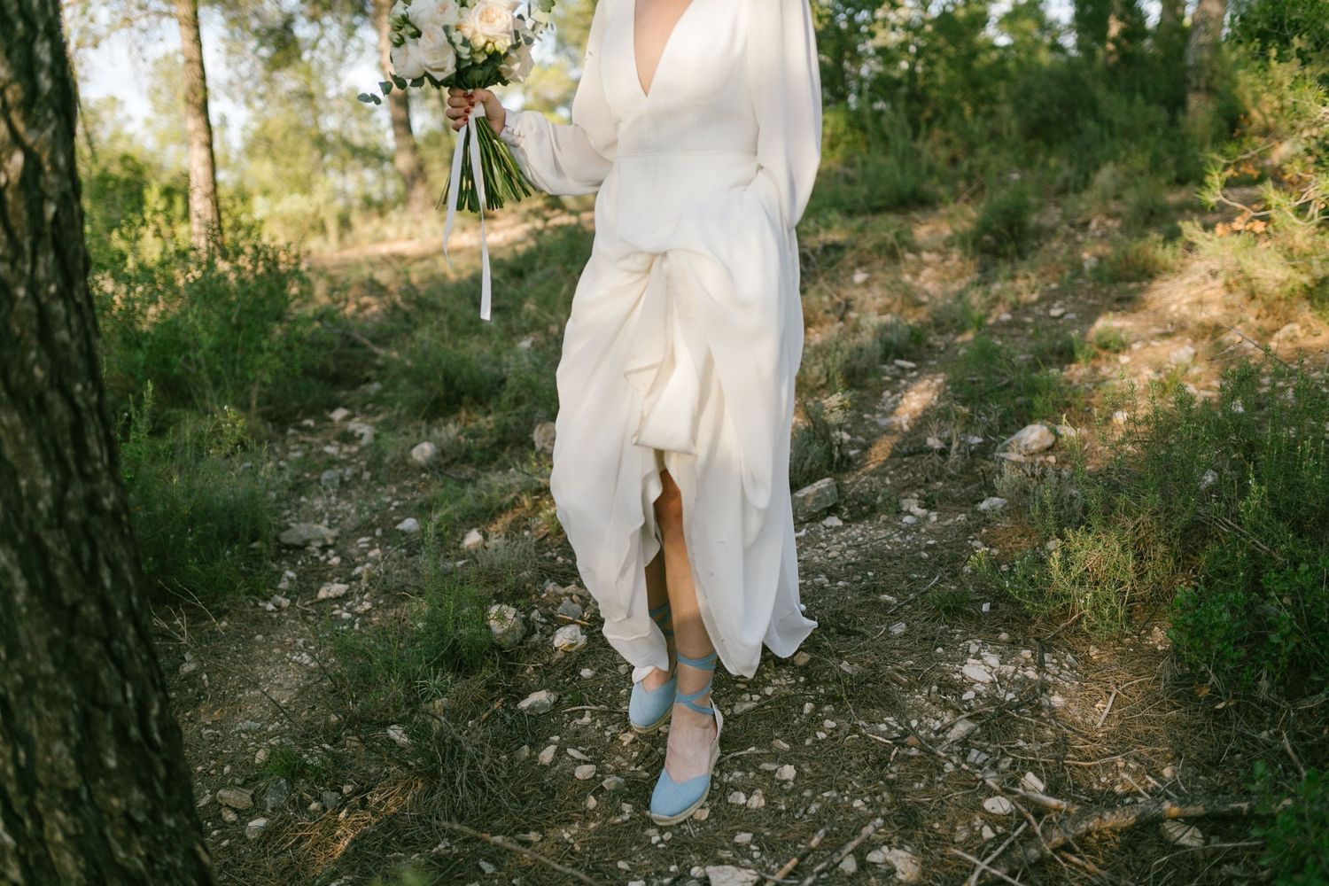 Close-up view of flowing white dress and blue shoes walking on a rugged outdoor path surrounded by greenery.