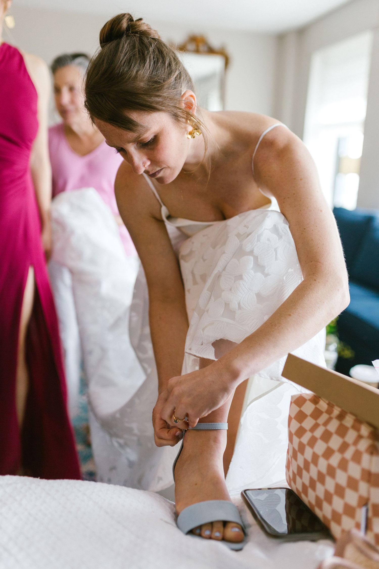 A woman in a white strapless wedding dress leans down to fasten her shoe while getting ready.