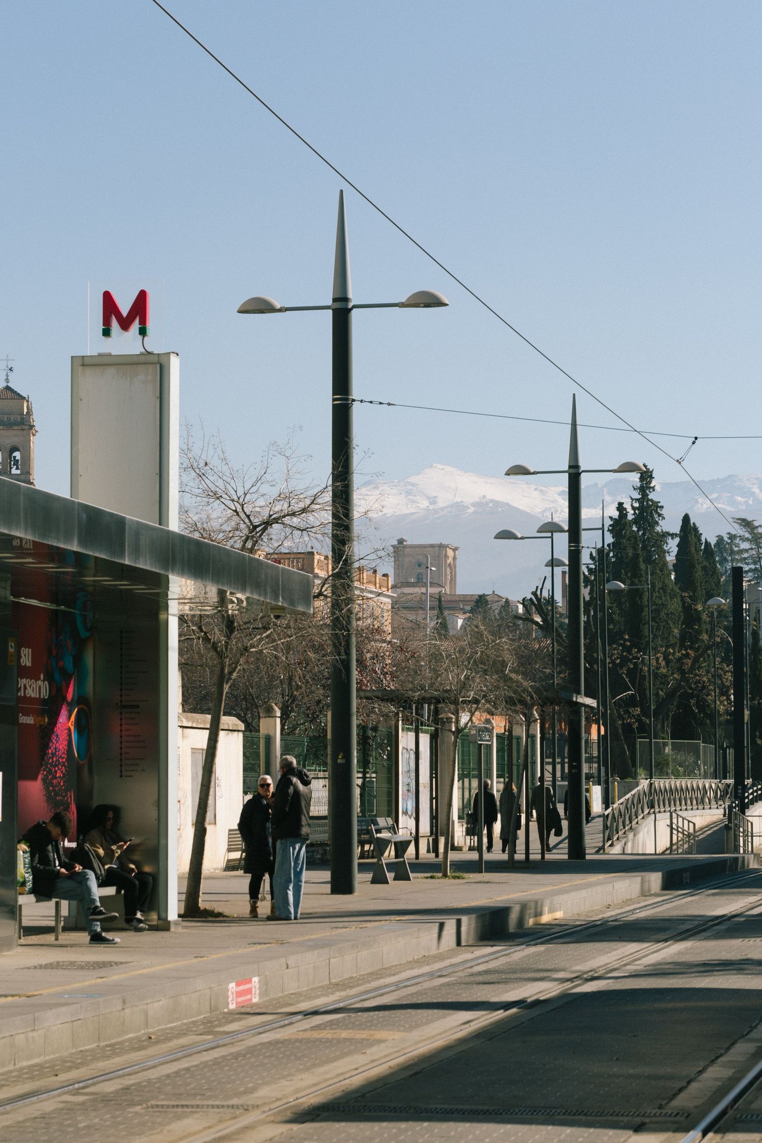Urban transit stop in Granada, Spain with metro entrance and streetlights along a city sidewalk beneath a clear blue sky.