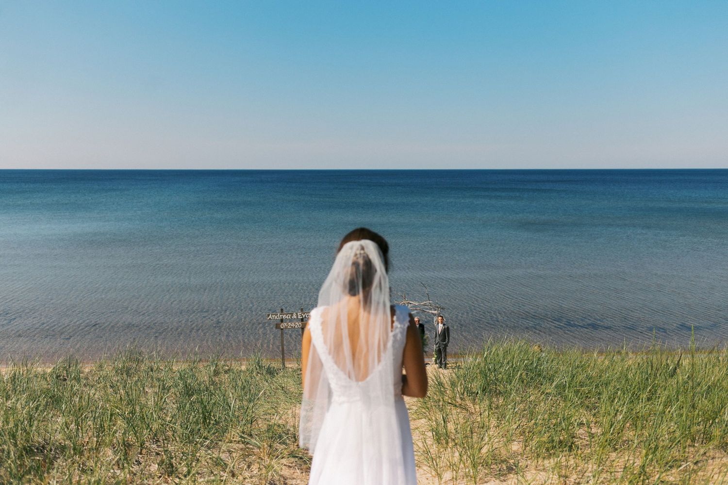 Rear view of a bride in white dress and veil overlooking a serene blue ocean on a clear day.