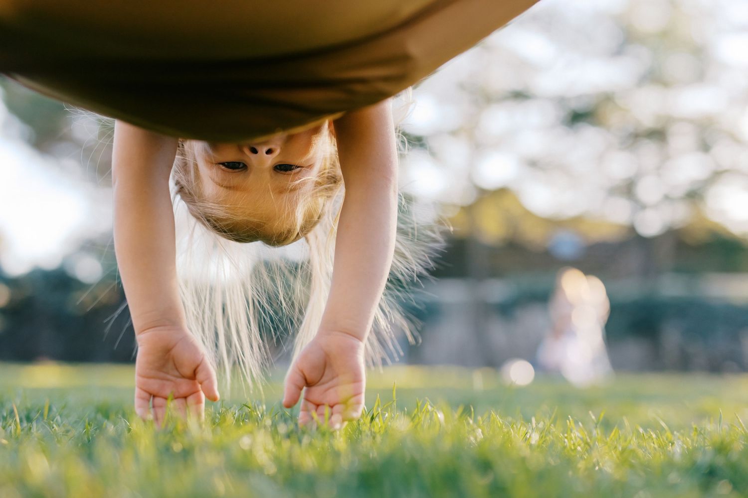 Young blonde girl bending over to touch green grass in a sunlit park.