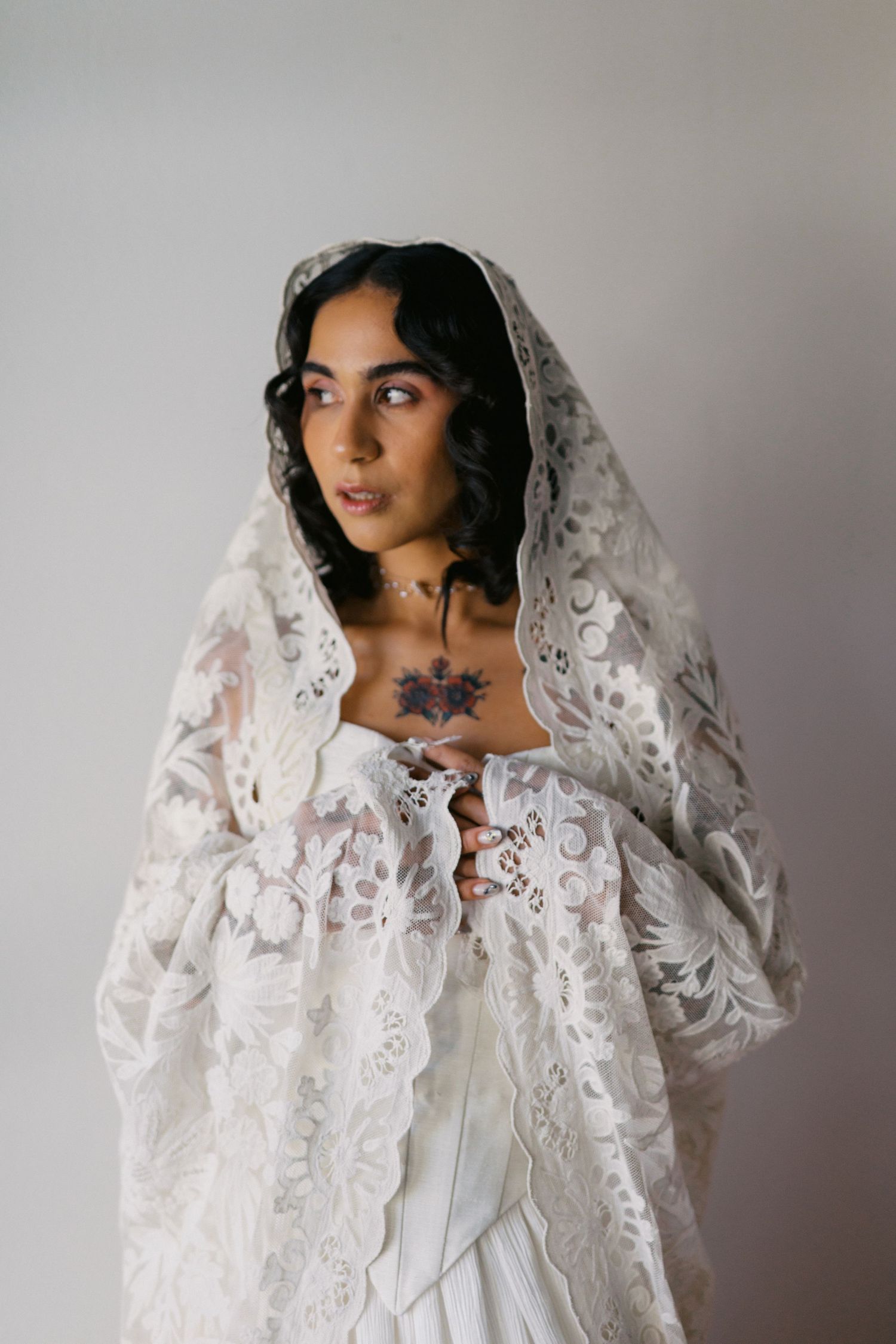 Close-up portrait of bride with chest tattoo wearing intricate white lace mantilla veil and wedding gown.