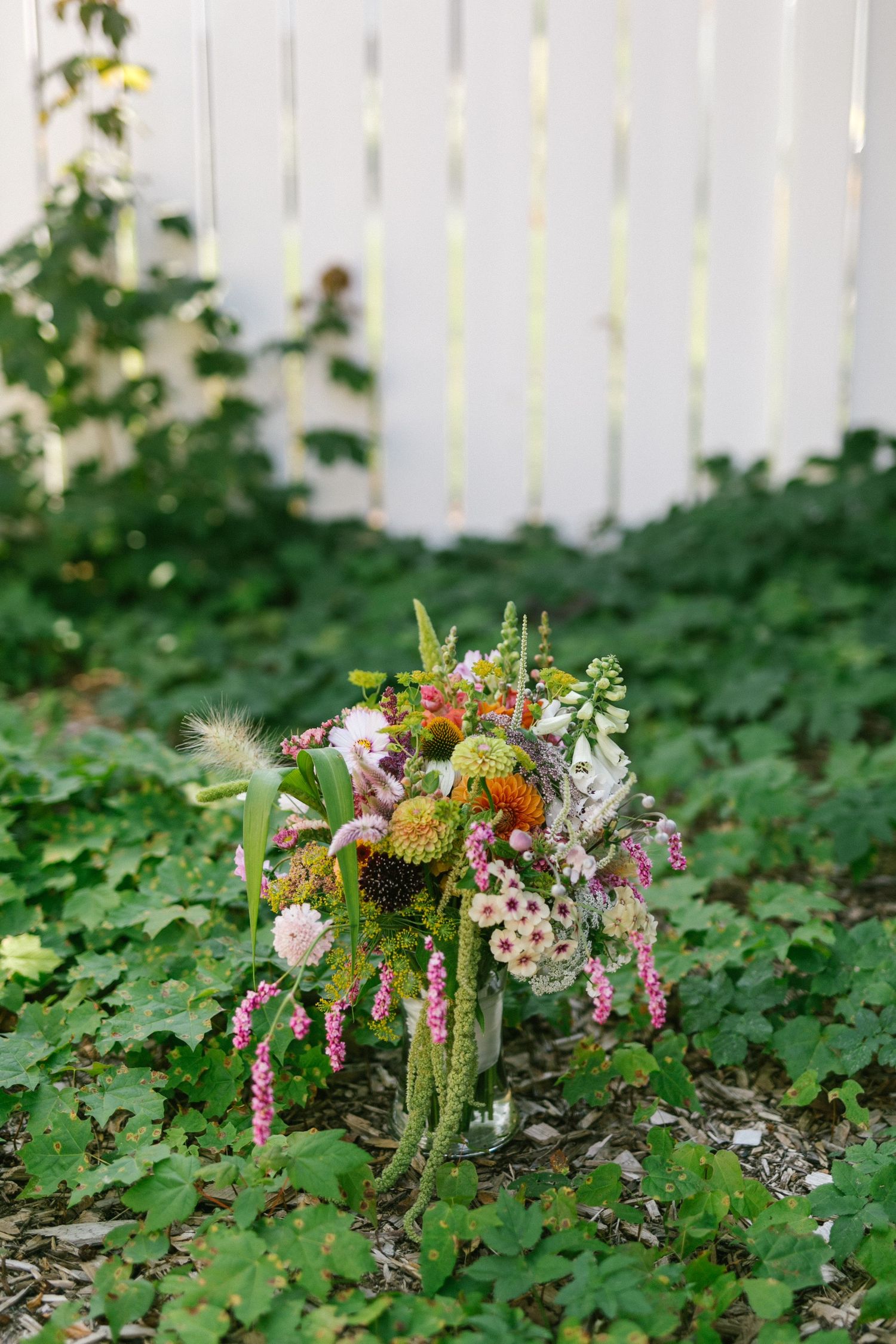 A colorful floral arrangement sits nestled among green foliage against a white fence in a garden setting.