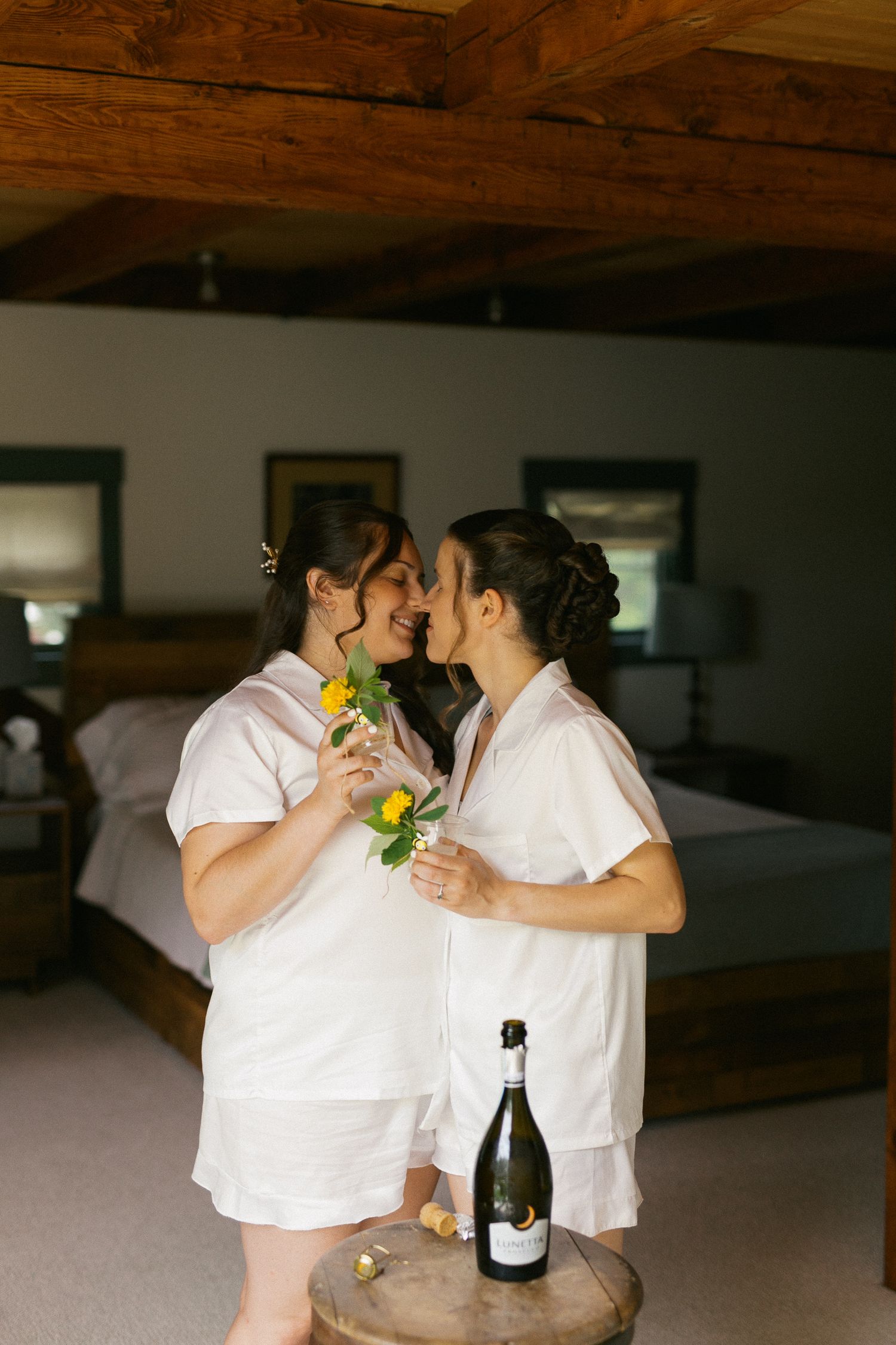 Two brides in white pajamas share a kiss holding yellow flowers with champagne bottle on rustic wood table.