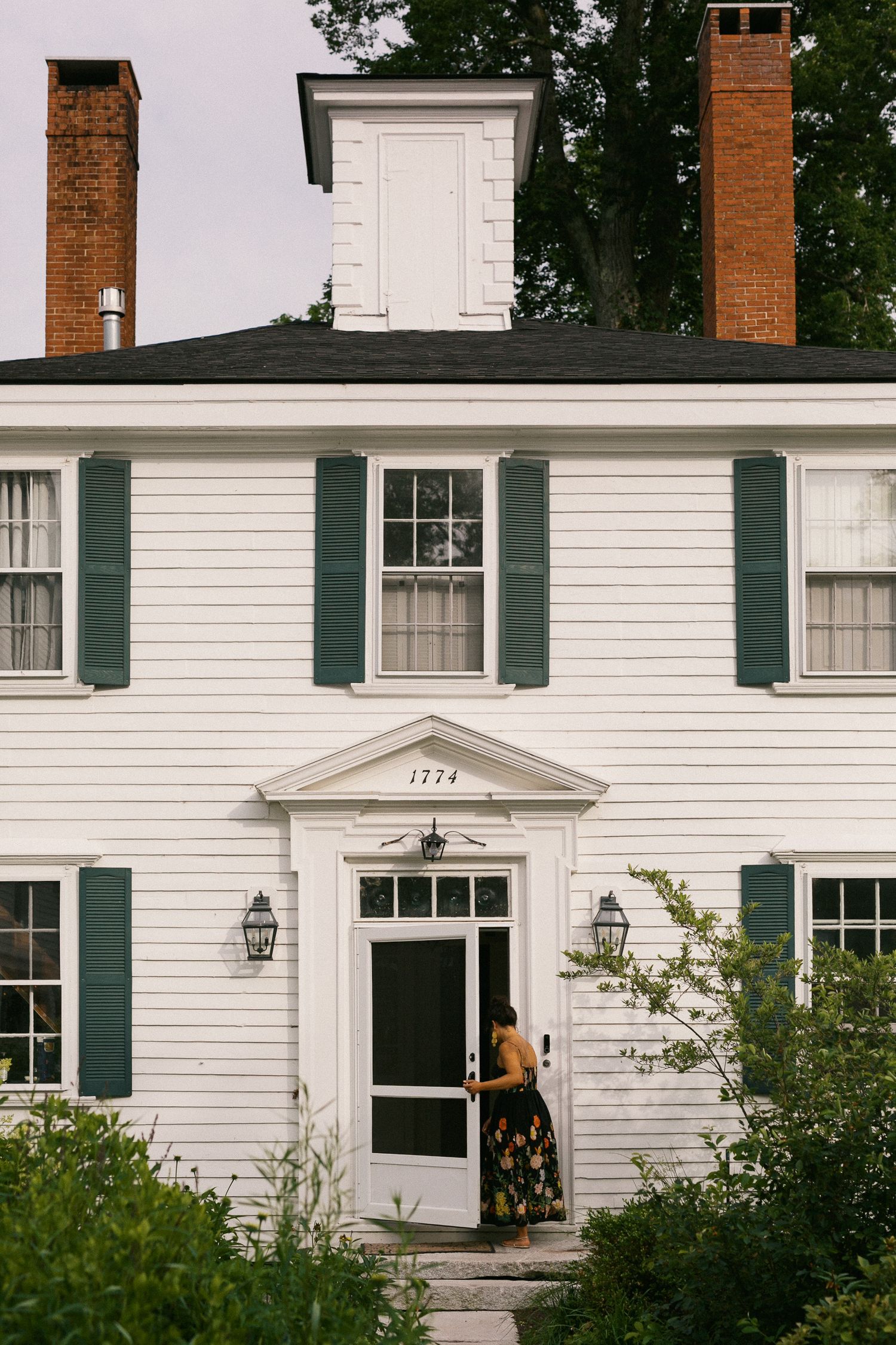 White colonial home with green shutters and brick chimneys, woman in floral dress entering front door.