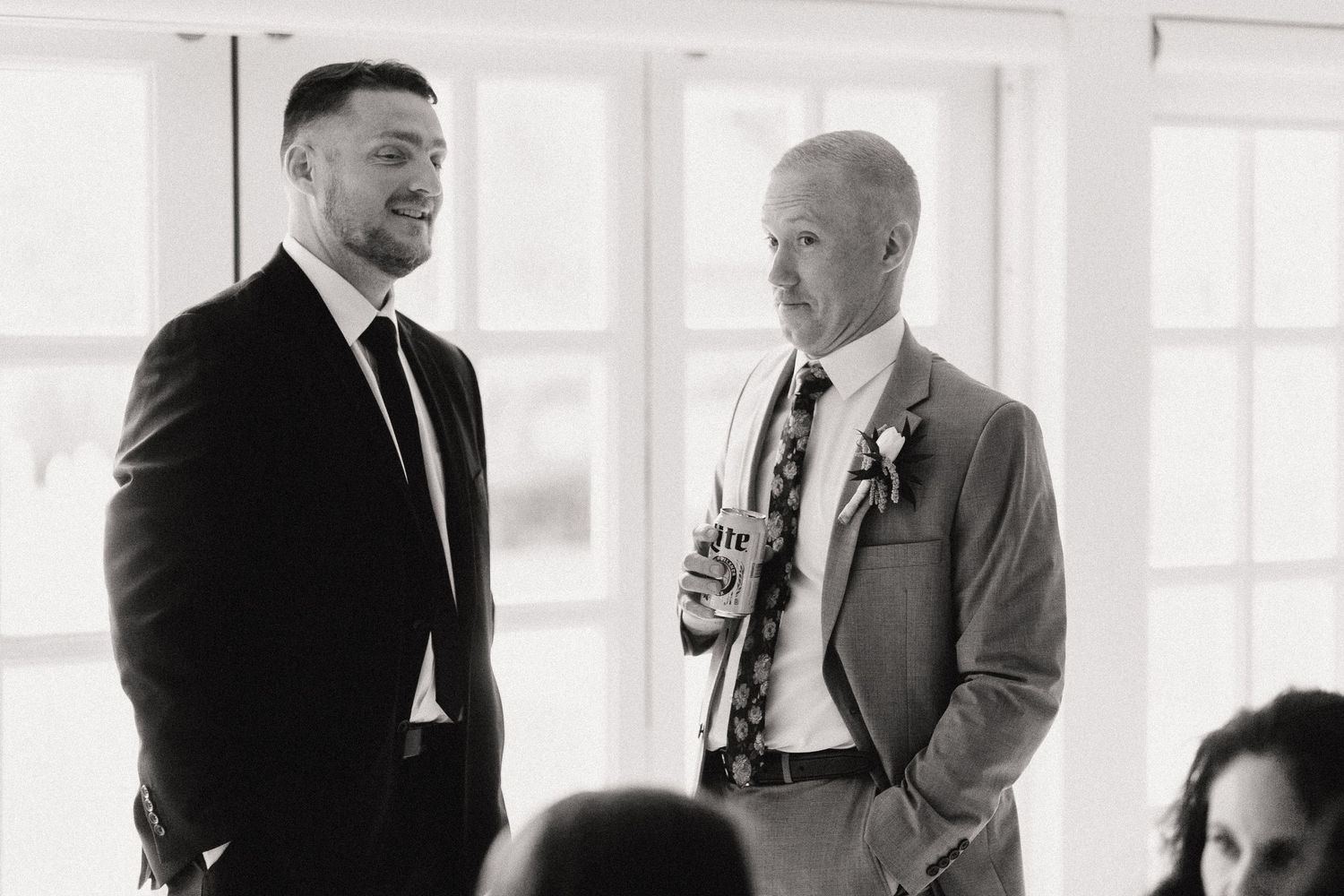 Two people in formal attire have an animated conversation in a bright room with large windows.