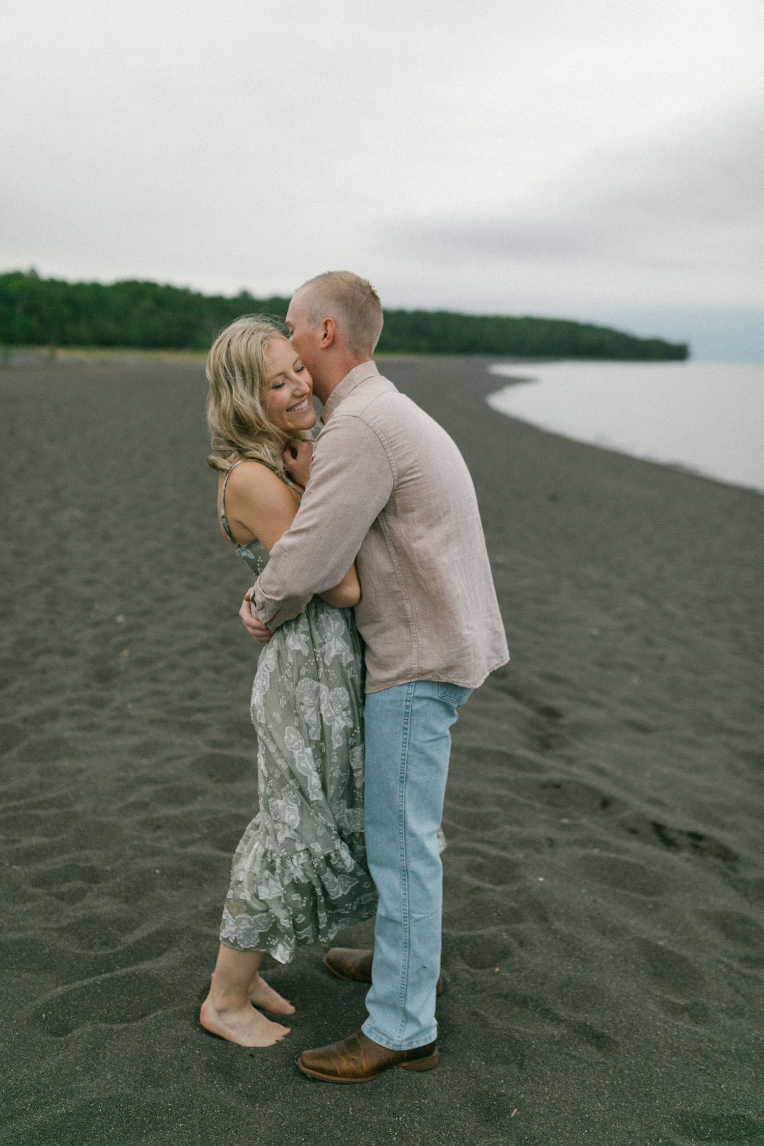 A romantic couple shares an intimate moment while standing barefoot together on a dark sandy beach.