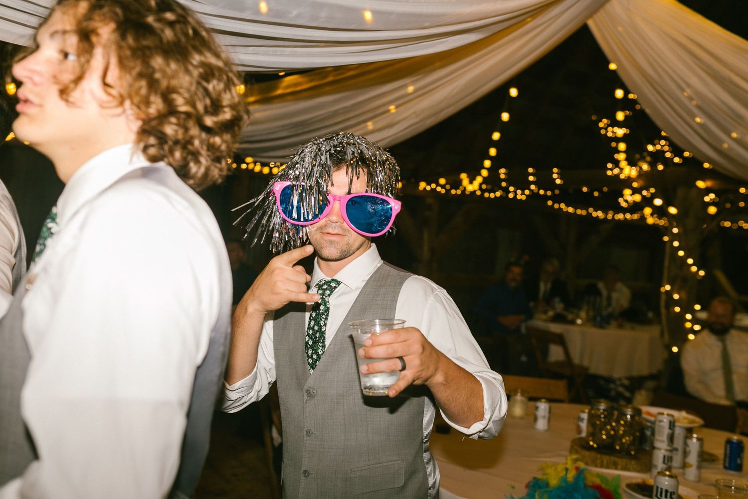 Party guest in vest and metallic star sunglasses holds drink on dance floor under draped tent with twinkling lights.