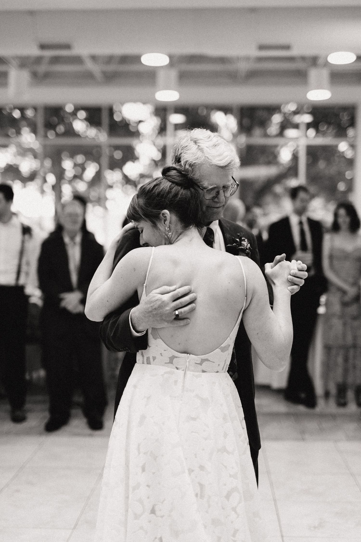 A couple shares an intimate dance in black and white at a wedding reception as guests look on from the background.