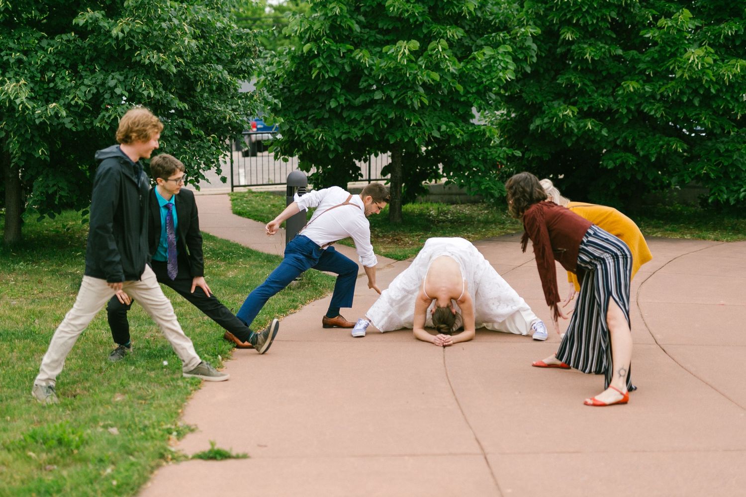 A group of people playfully pose on a concrete path surrounded by green trees and grass.