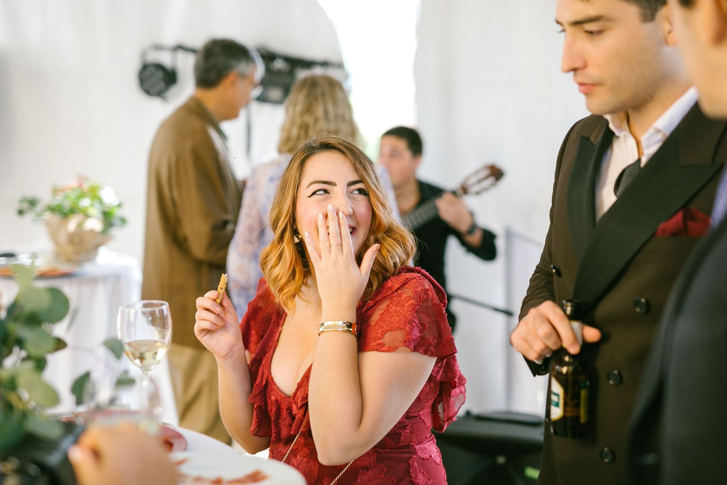 A woman in a red dress reacts with surprise while someone in a suit stands nearby at an outdoor wedding.