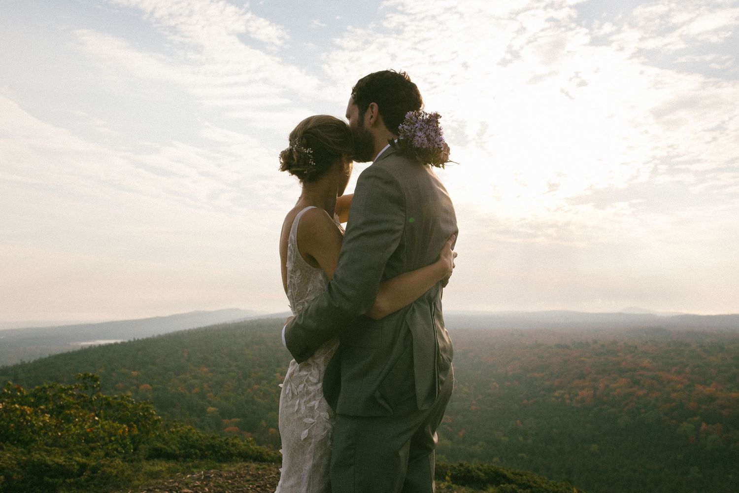 A couple shares a romantic embrace at sunset overlooking a mountain valley landscape in the warm evening light.