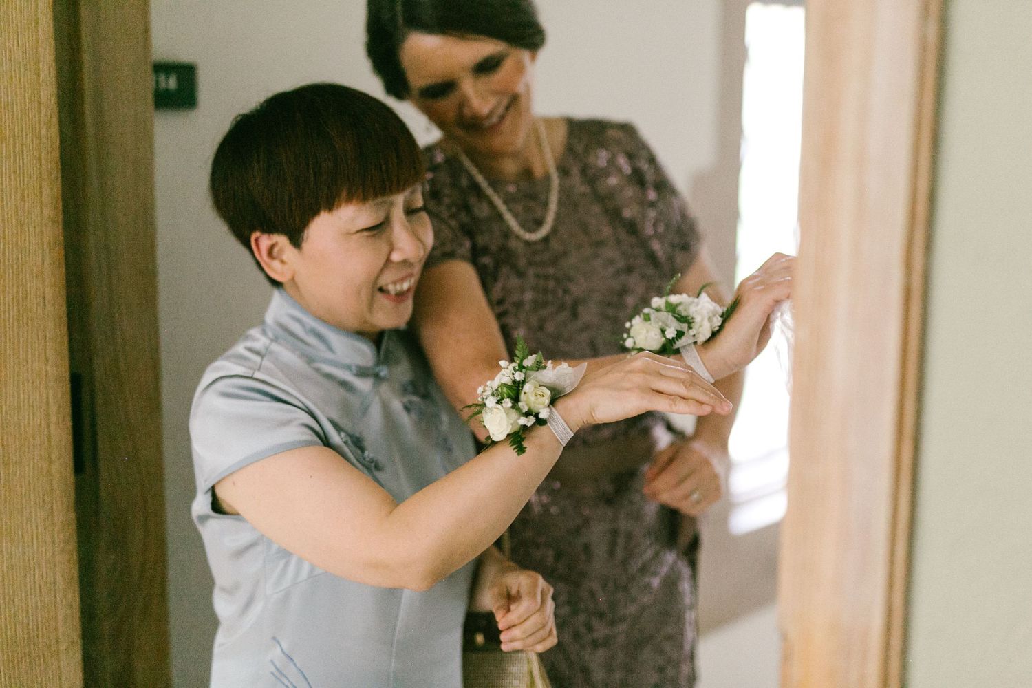 Young boy helping pin a floral corsage onto a woman's wrist during wedding preparations indoors.