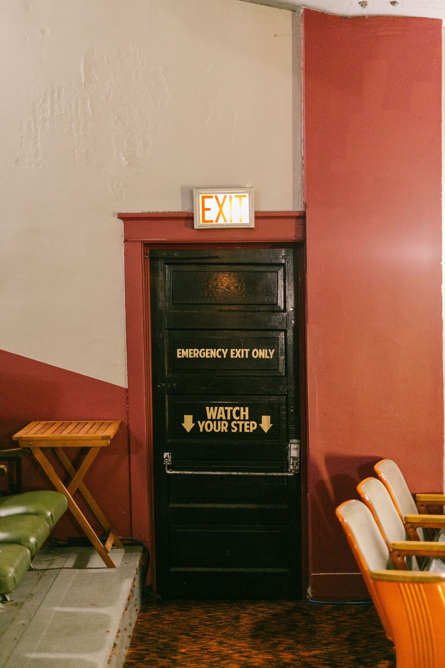 Dark wooden emergency exit door with warning signs mounted in a red walled interior space with vintage seating.