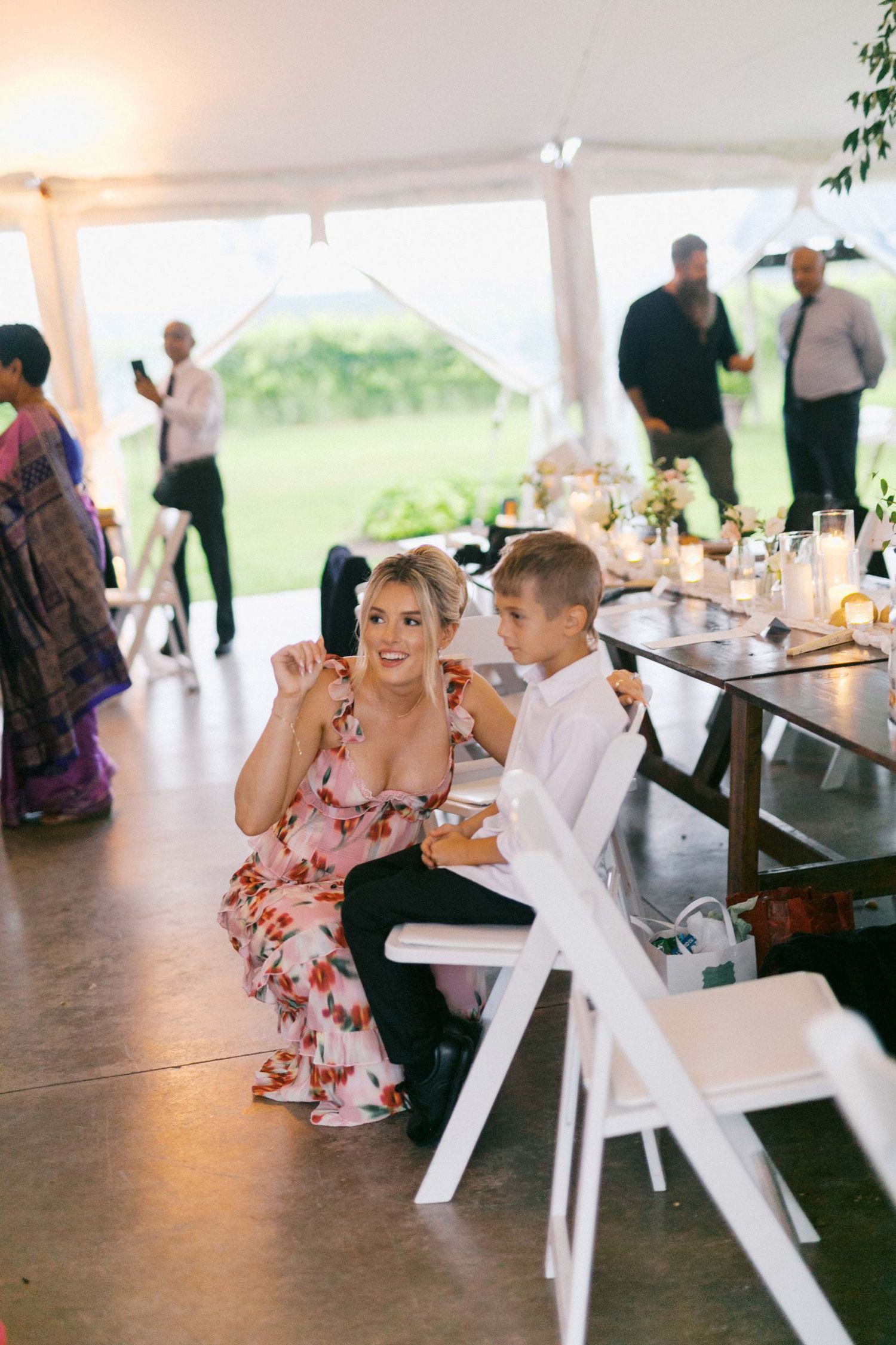 A couple sits together at white picnic tables under an outdoor tent during a wedding reception.