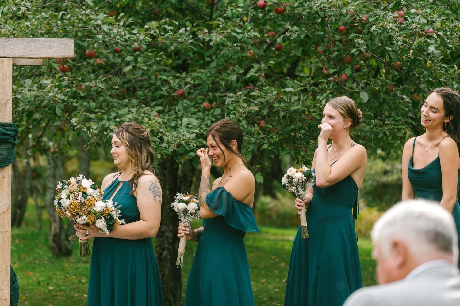 Bridesmaids in emerald green dresses hold bouquets during an outdoor wedding ceremony.