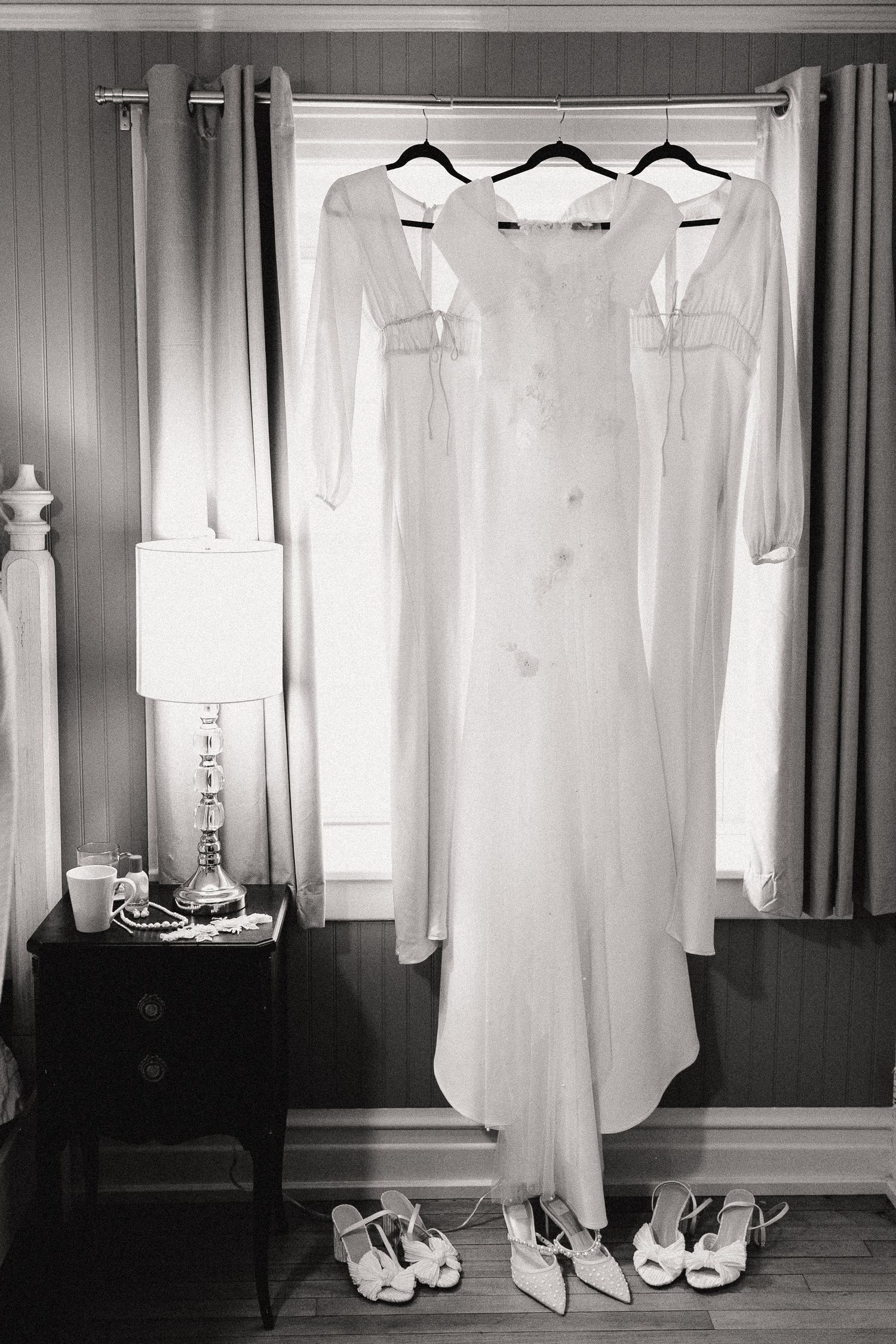 Three white dresses hang on display with shoes lined up beneath them and a lamp on a side table.