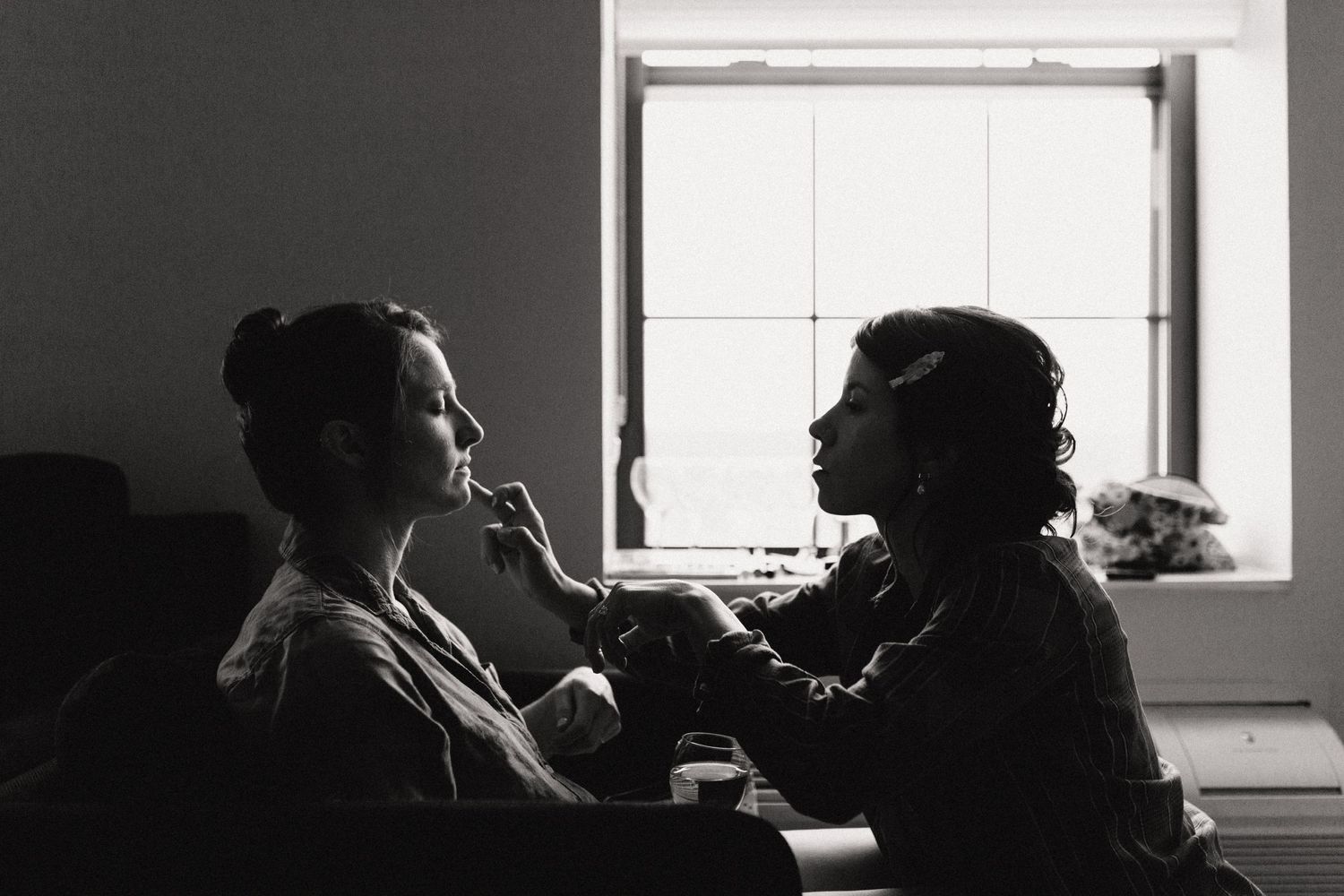 A black and white photograph shows two silhouetted figures seated by a window during what appears to be a makeup application.