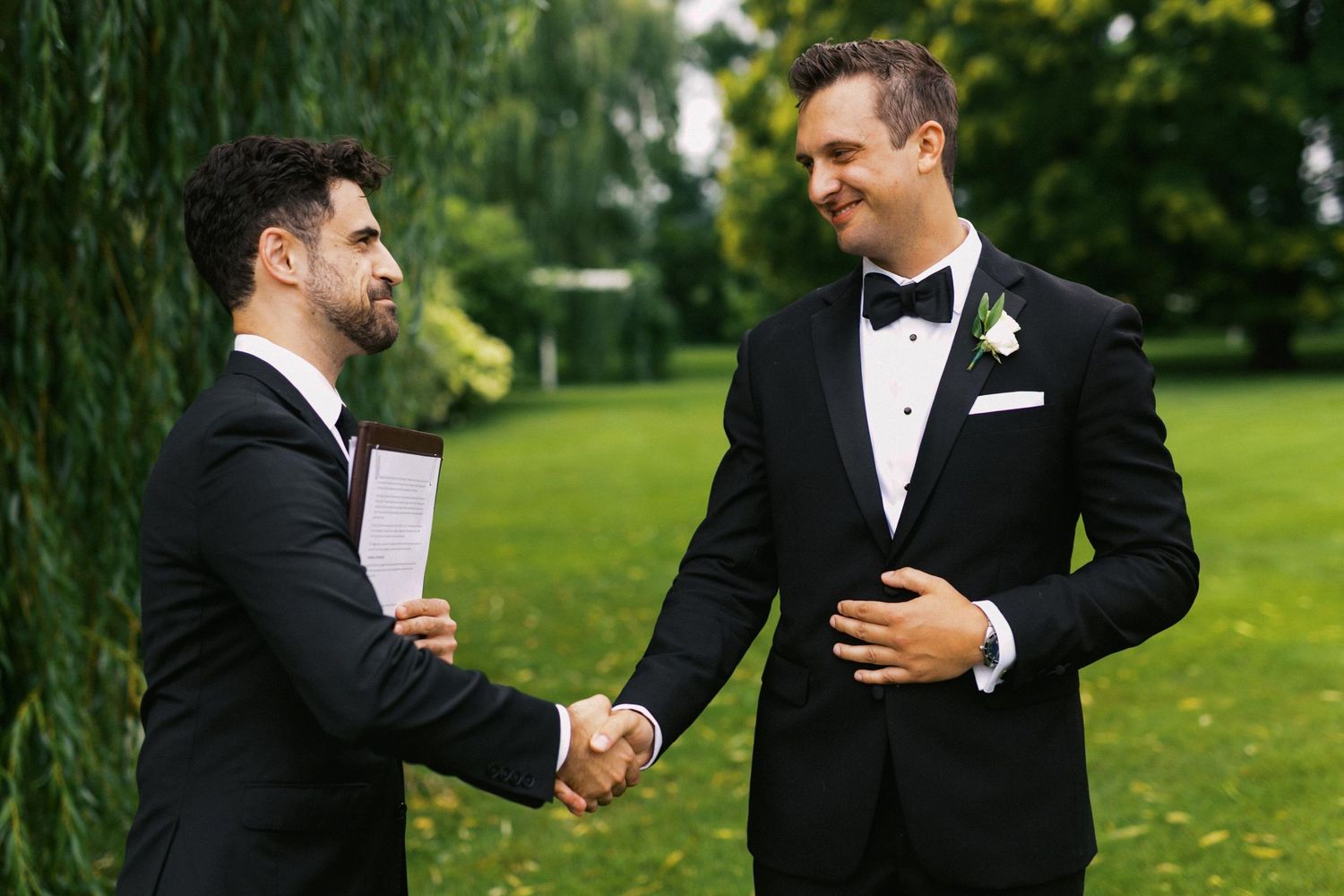 A groom and his friend the officiant in formal black tuxedos shake hands and smile while standing on a lush green lawn.