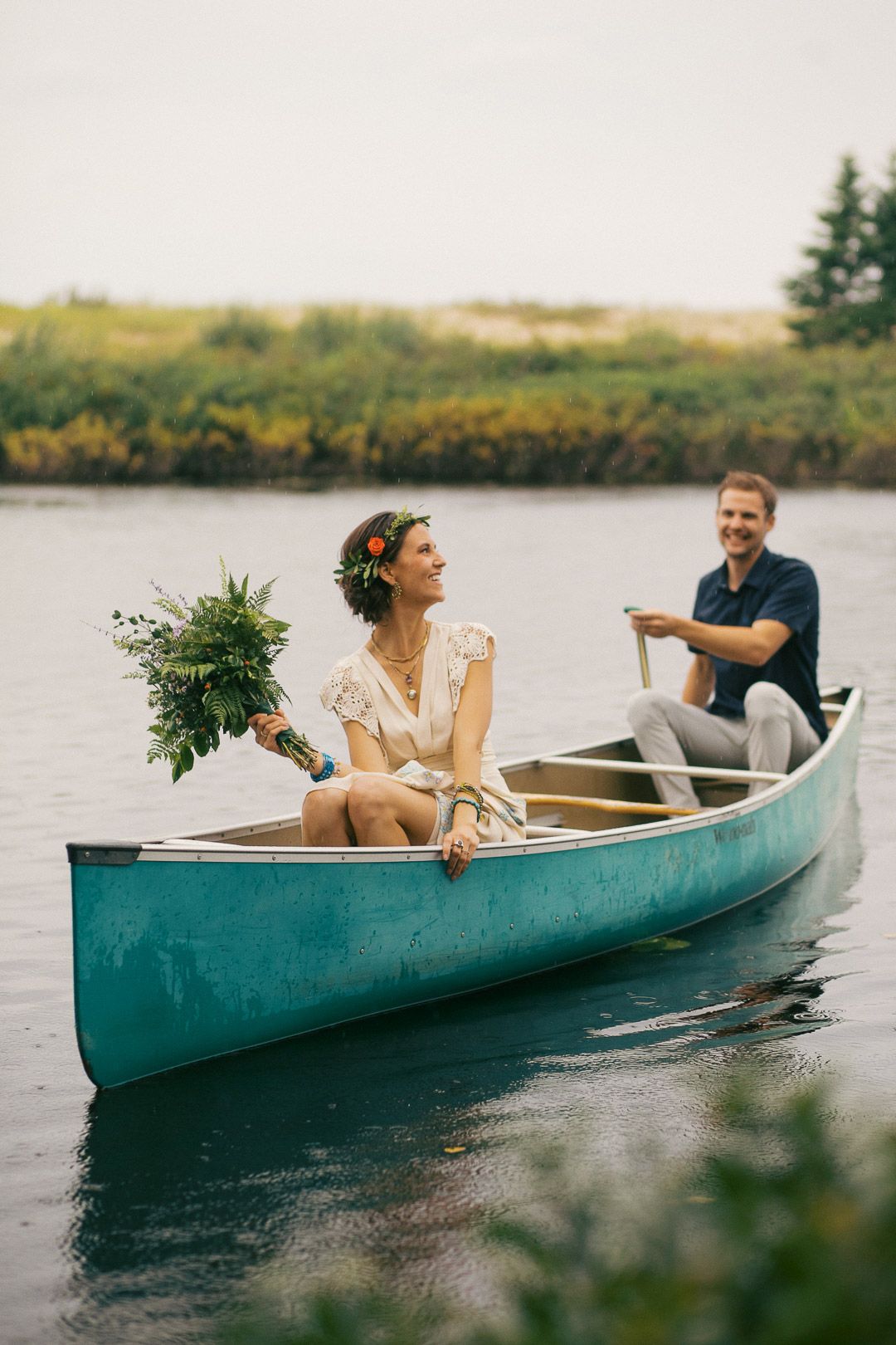 Couple in a teal canoe on a calm lake, bride holding a bouquet with floral crown, groom paddling in background.