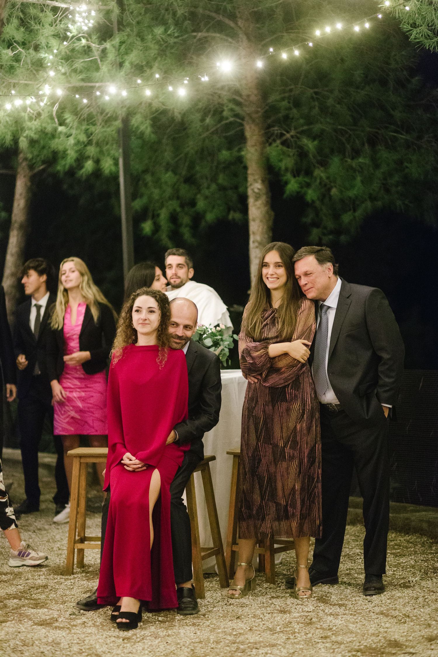 A group of wedding guests gather under string lights at an outdoor evening reception.