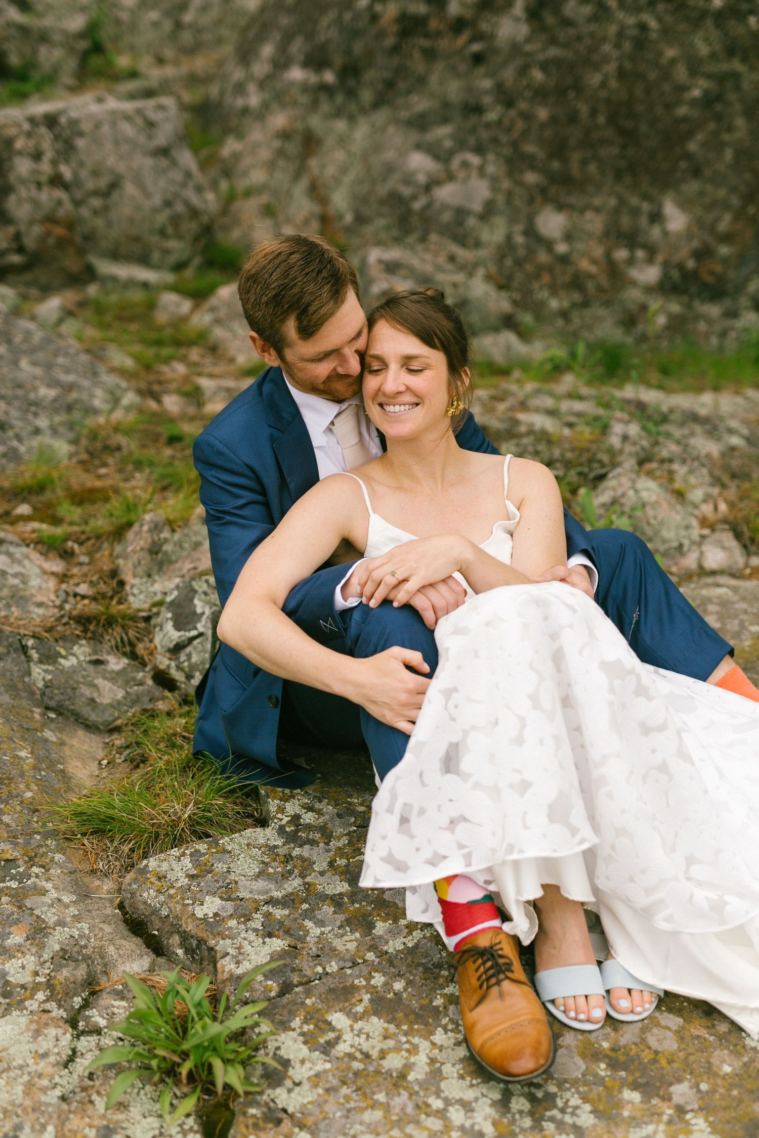 A couple in wedding attire sits intimately on rocky terrain, wearing a blue suit and white lace dress with casual footwear.