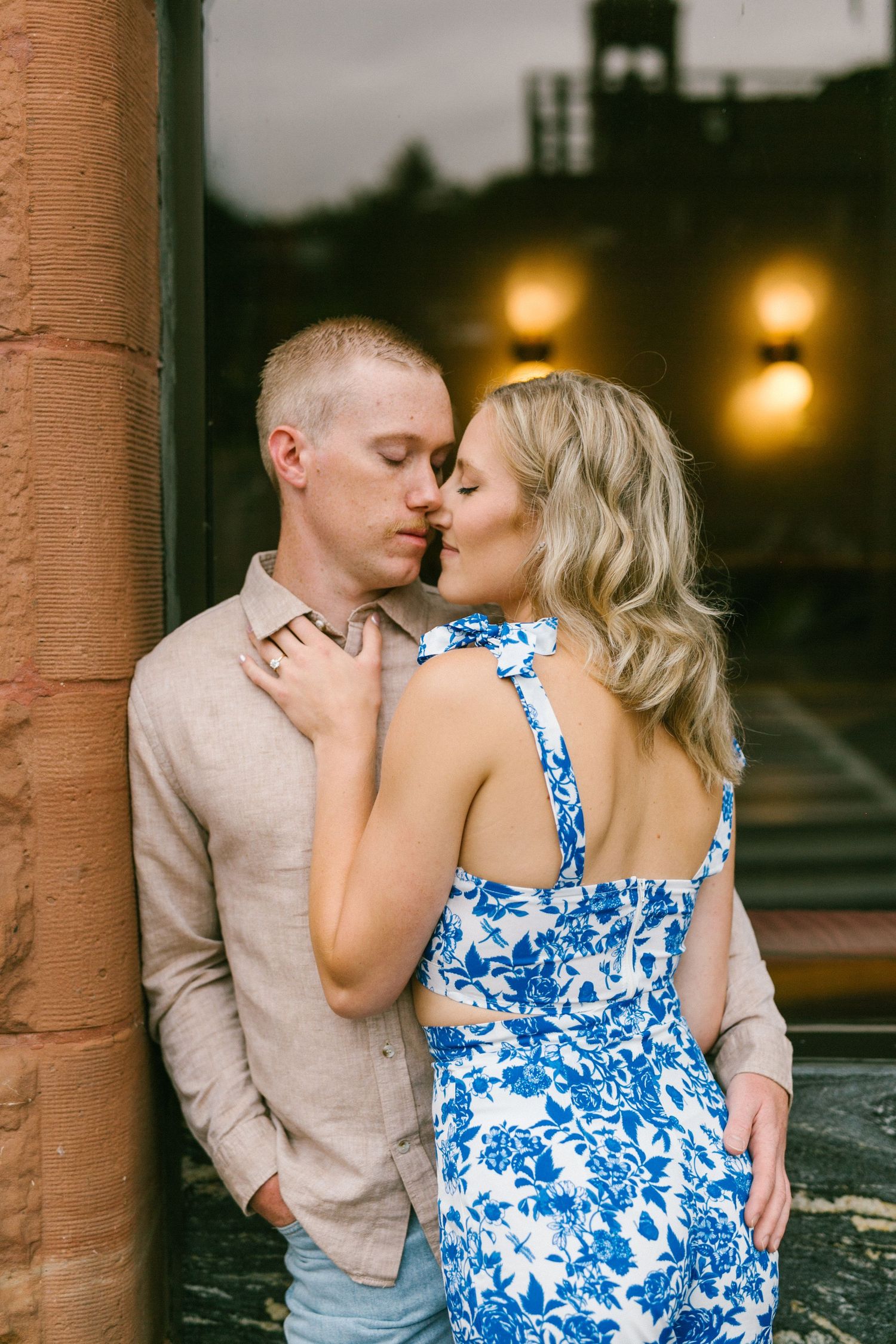 Romantic couple embraces near historic building at dusk with glowing lights in background.