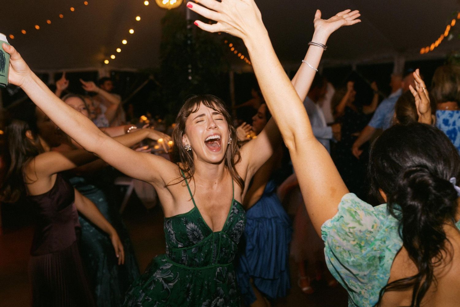 People dancing and singing with their arms raised at a lively wedding dance party with string lights overhead.