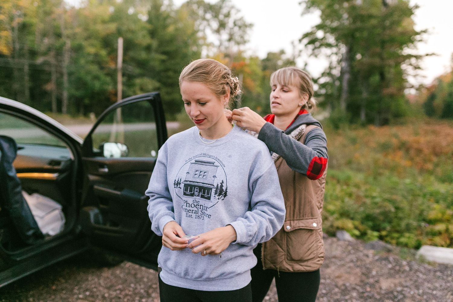 Two people stand near a parked car outdoors, one styling the other's hair in a casual autumn setting.