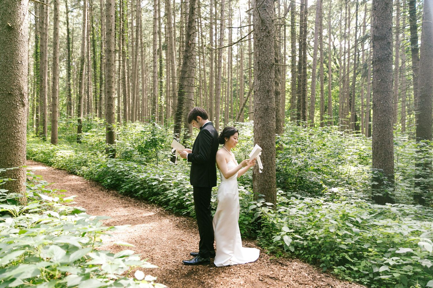 Bride and groom stand back-to-back reading vows on a forest path surrounded by tall trees and lush greenery.