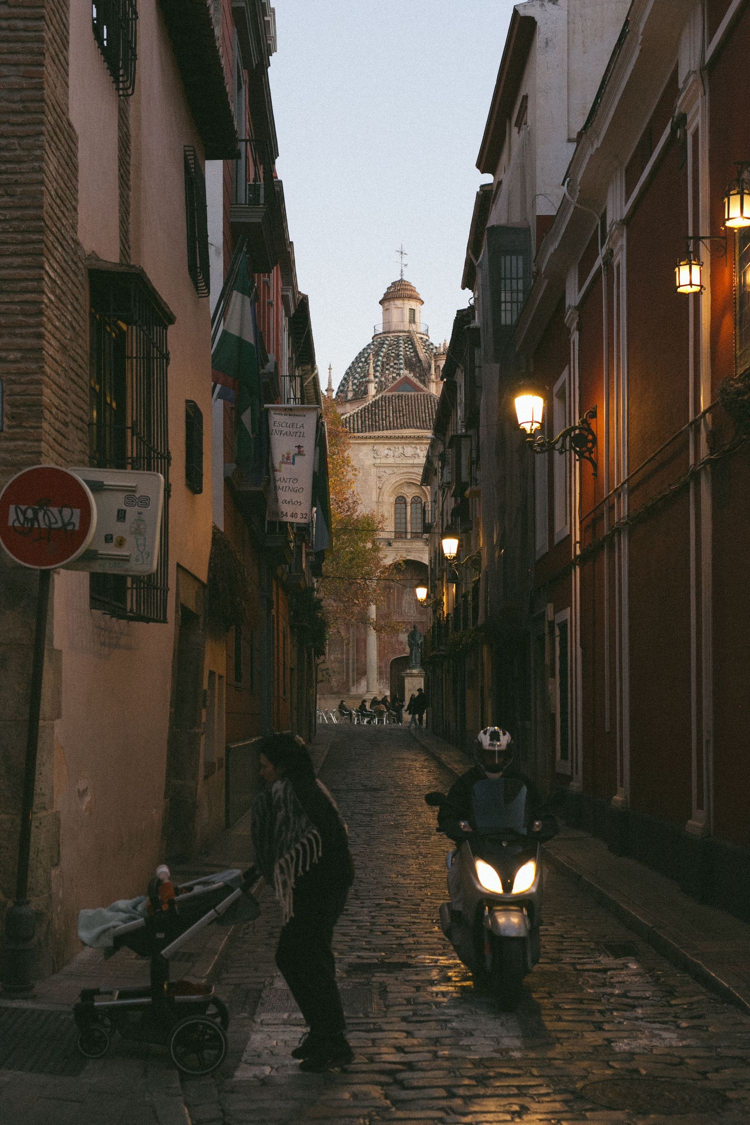Narrow cobblestone street in an old European city with a church dome visible in the distance and warm street lights.