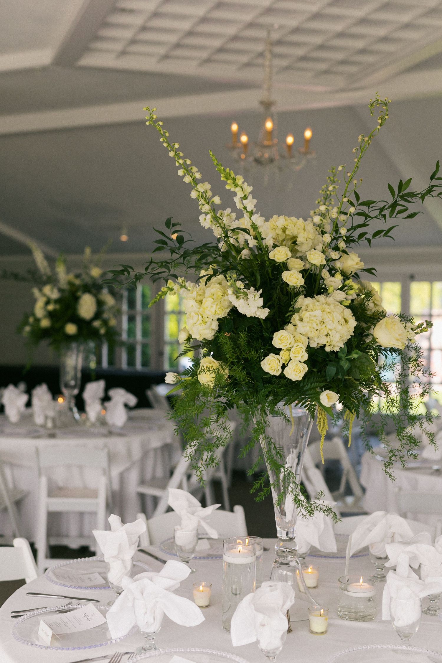 An elegant white wedding reception setup with tall floral centerpieces and crystal chandeliers.
