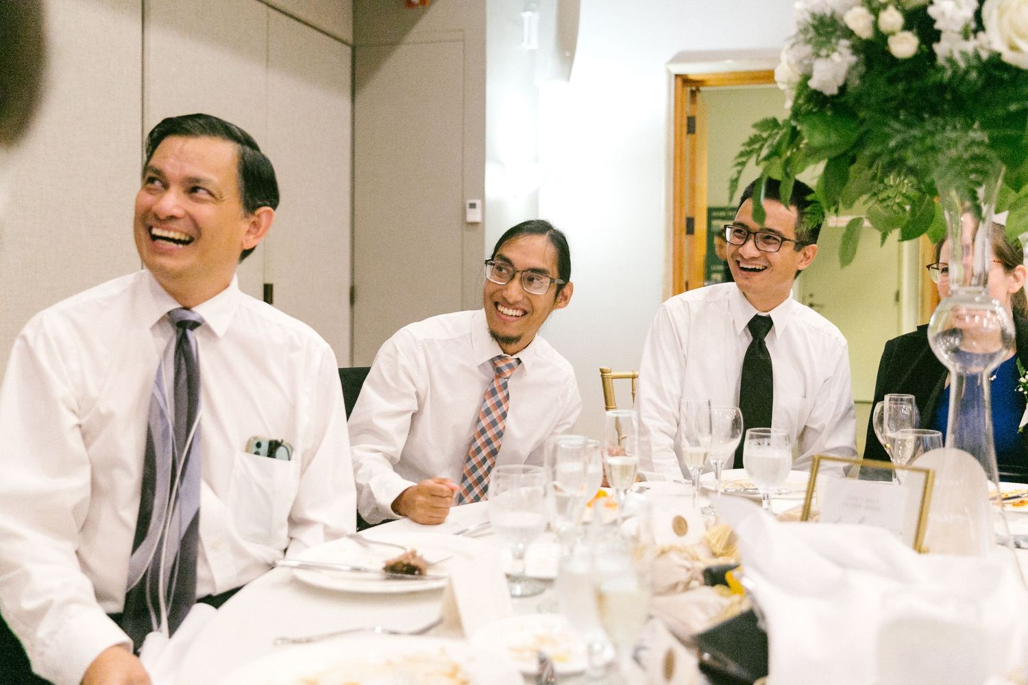 Three men in dress shirts and ties laughing joyfully at a formal wedding reception dinner table.