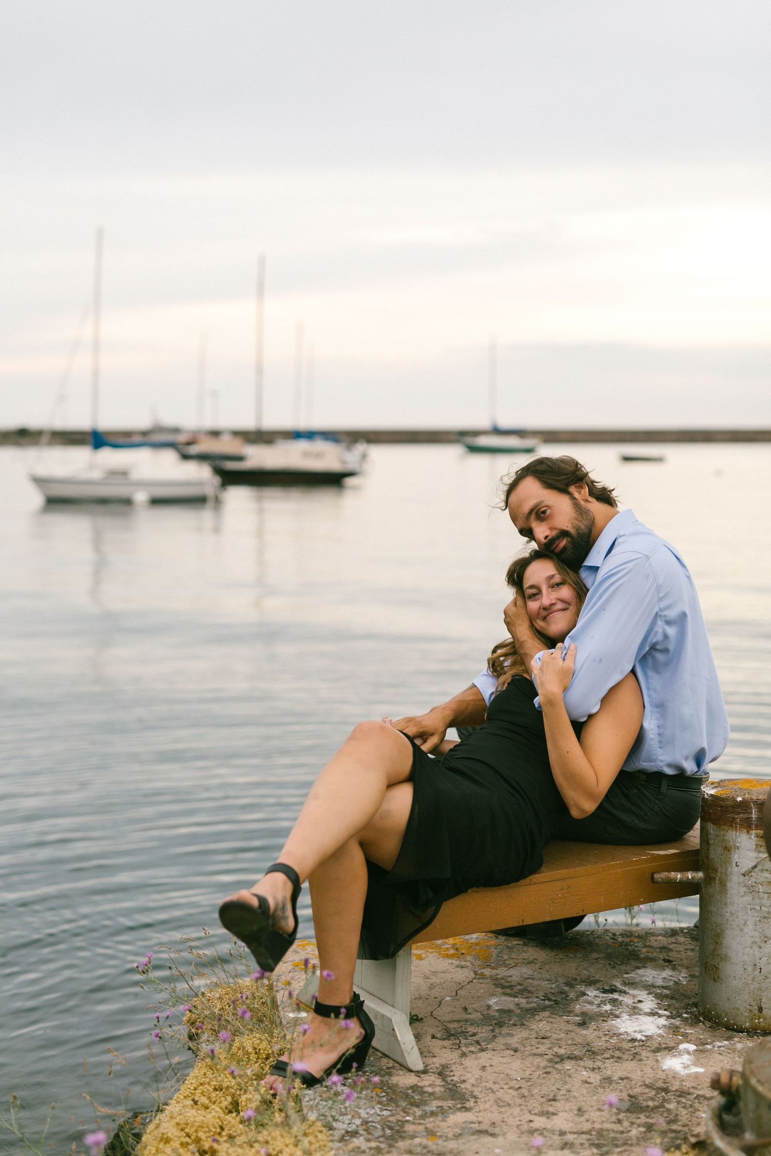 A romantic couple shares an intimate moment while sitting on a dock with sailboats in the background.