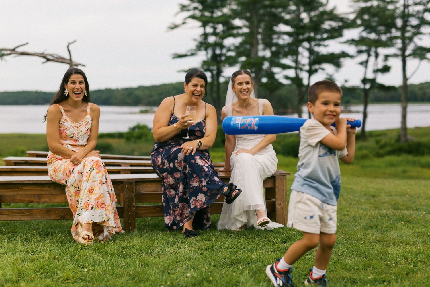 Three women sit on wooden benches outdoors near a river while a young boy hits balls with a blue baseball bat.