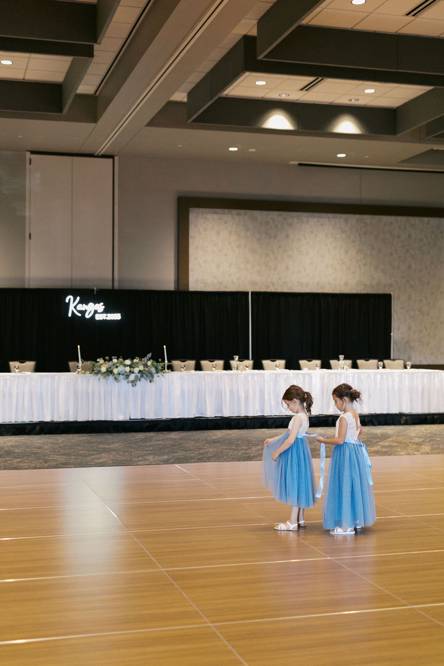 A panoramic sequence of two figures in blue dresses walking across an empty ballroom with white drapes in the background.