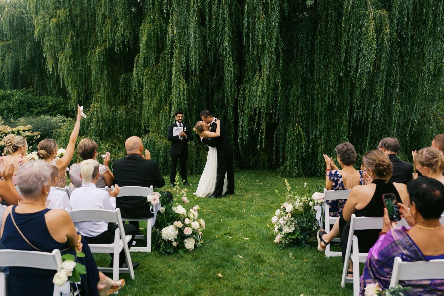 A bride and groom share their first kiss as husband and wife under a magnificent willow tree as their guests cheer around them.
