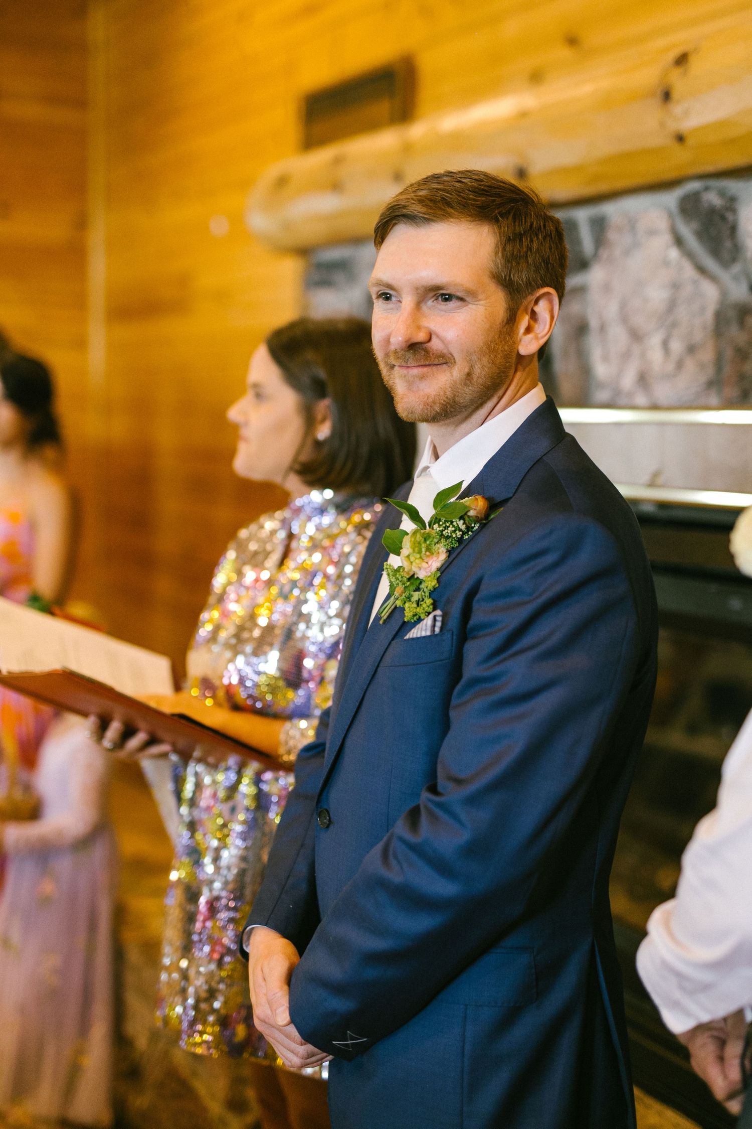 The groom in a navy suit with boutonniere stands during a rustic wooden cabin wedding ceremony.
