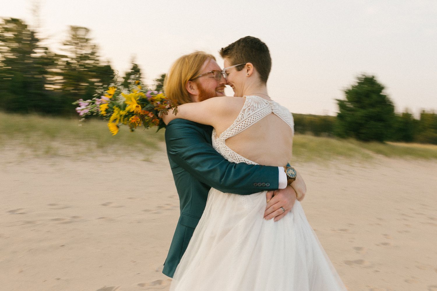 A couple embraces on a sandy beach at sunset with pine trees in the background.
