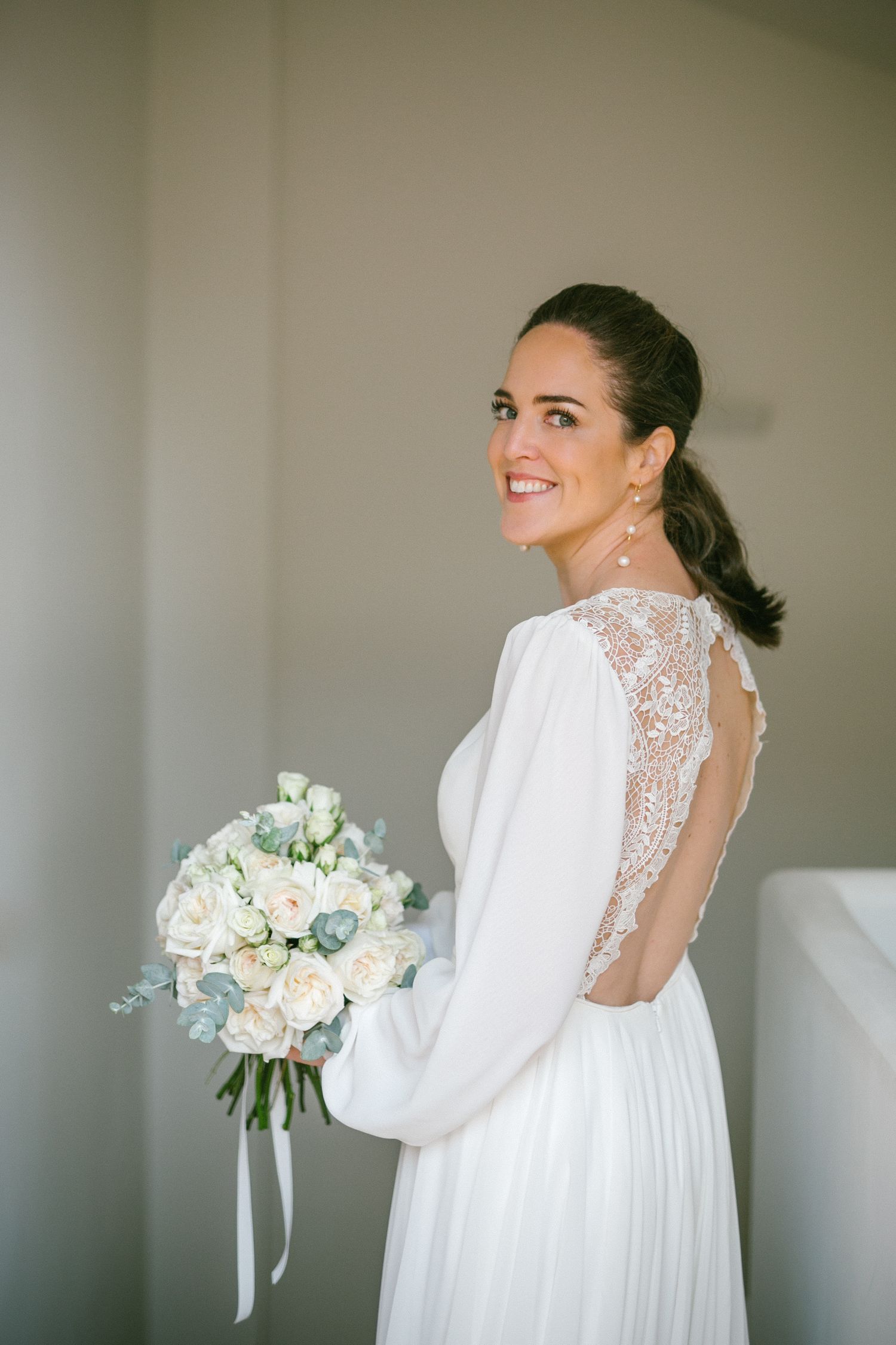 Elegante novia con vestido blanco de manga larga con detalle de espalda abierta de encaje sostiene romántico ramo de flores blancas y rubor.