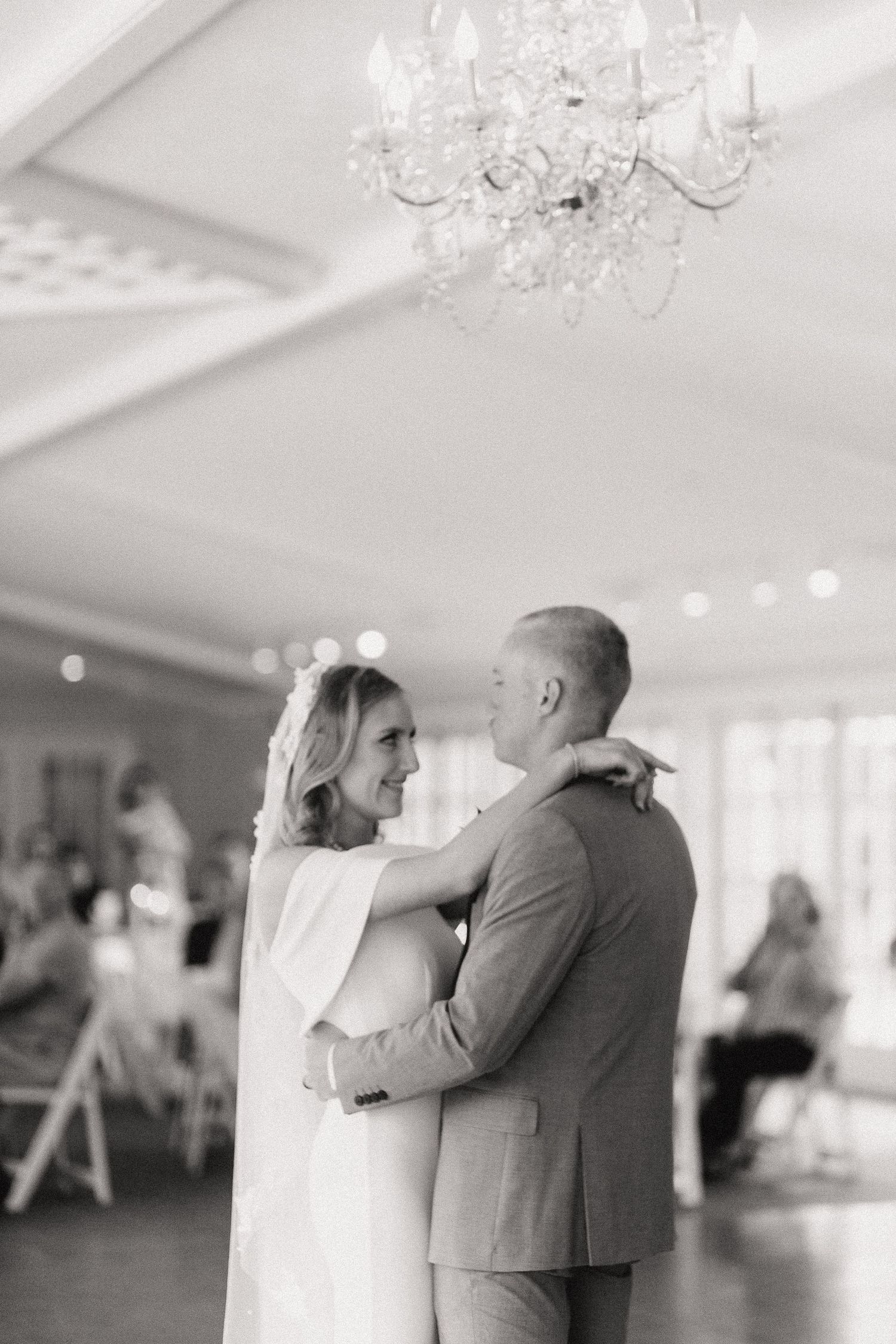A newlywed couple dances together beneath elegant crystal chandeliers in a bright reception hall.