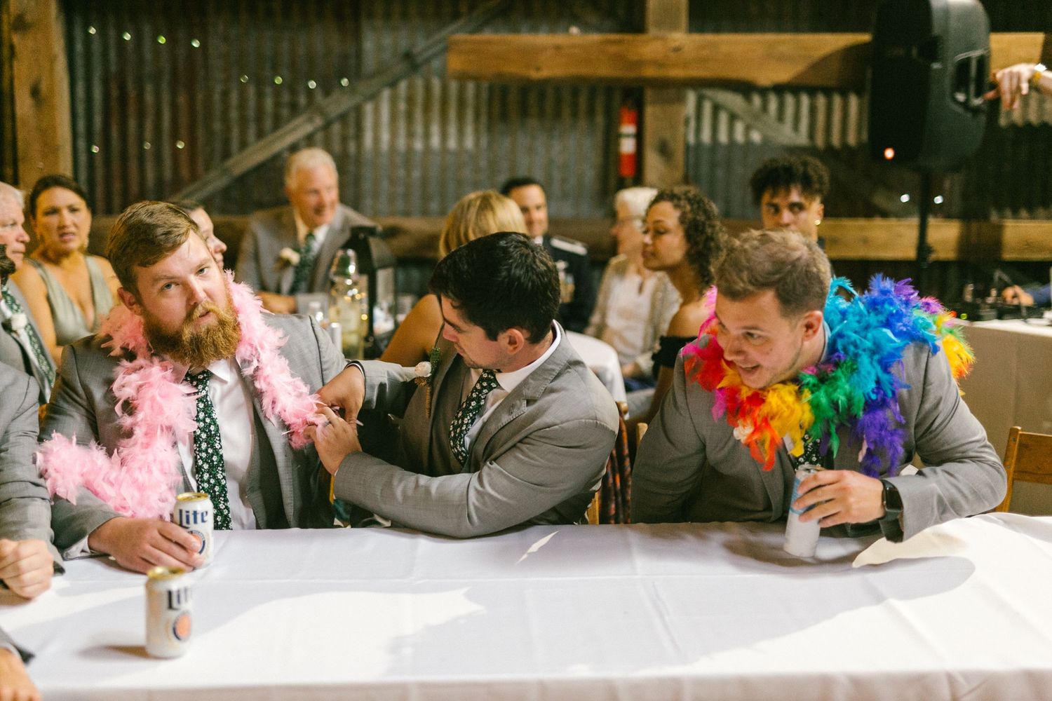 Celebration scene at a party showing people and groomsmen gathered around tables with colorful party decorations and feather boas.