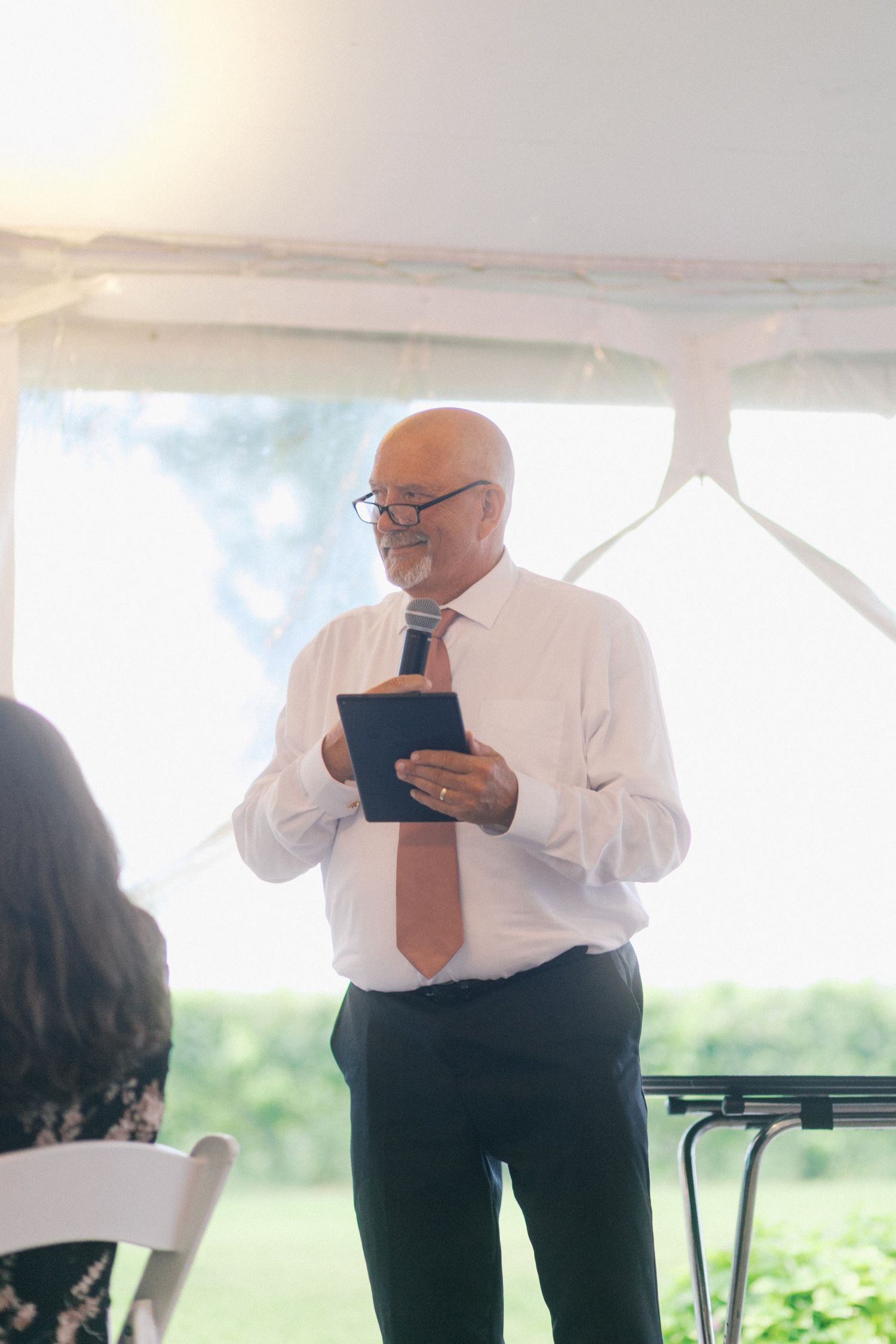 A person in formal attire with a white shirt and orange tie stands under a tent holding a tablet during an outdoor event.