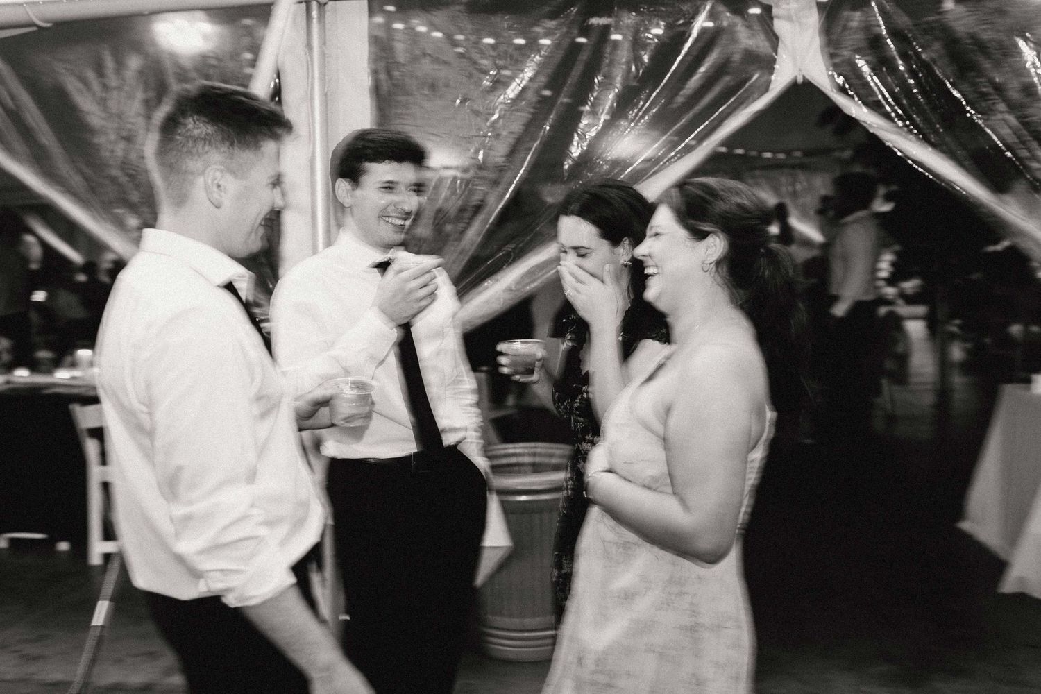 Black and white photo of people dancing and laughing at a wedding reception under string lights and decorative draping.