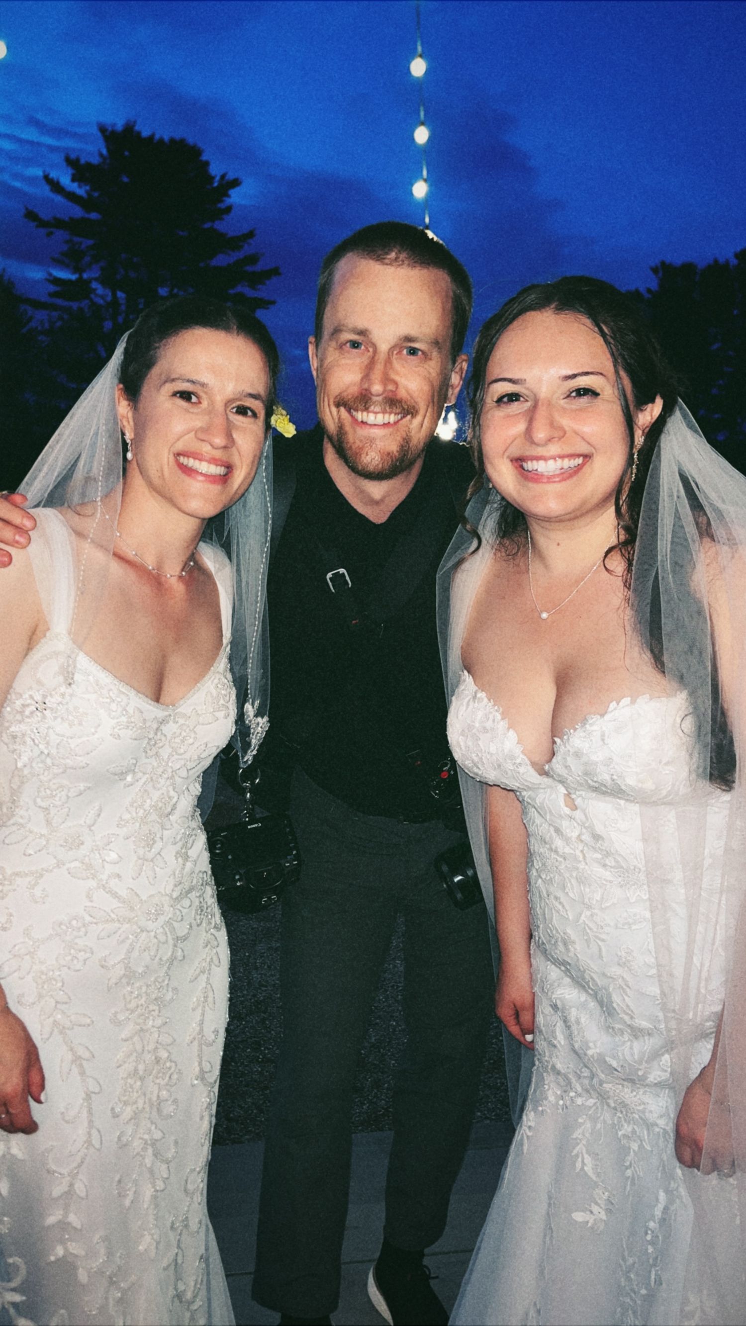 Two brides in white wedding gowns and veils pose smiling with Dylan of Miles North Creative . Co at an outdoor evening wedding celebration.