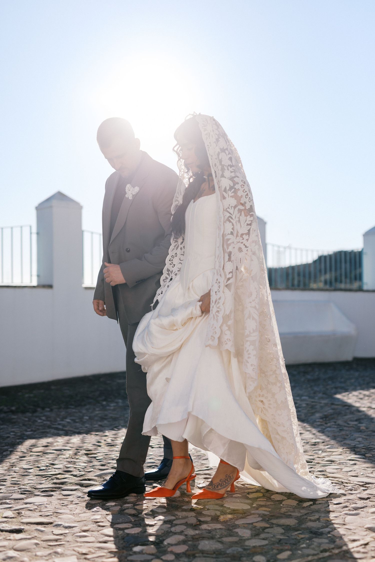 Backlit silhouette of bride in lace veil and orange heels with groom on cobblestone rooftop terrace.