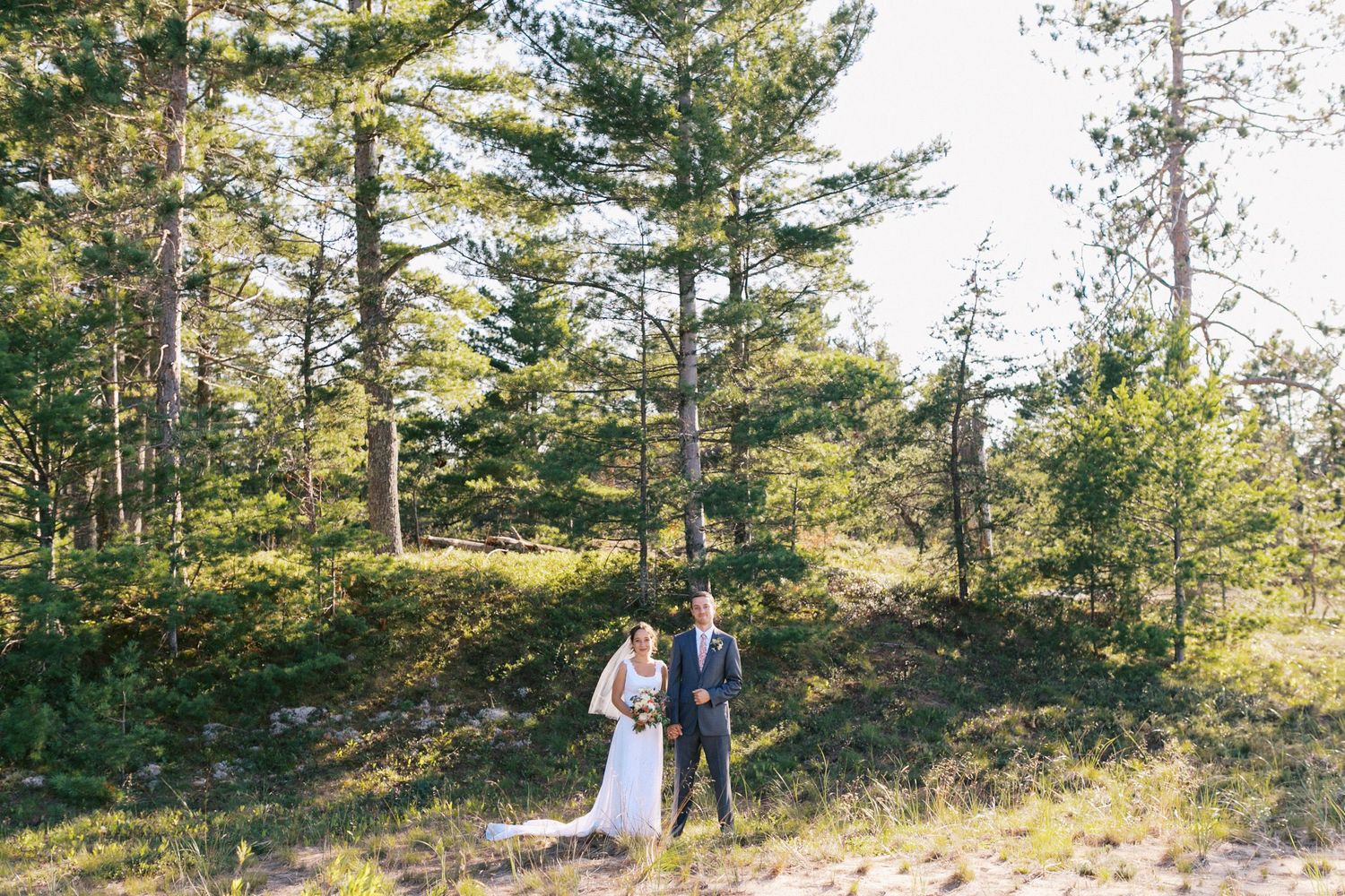 Bride and groom stand together among tall pine trees on sandy dunes in natural setting.