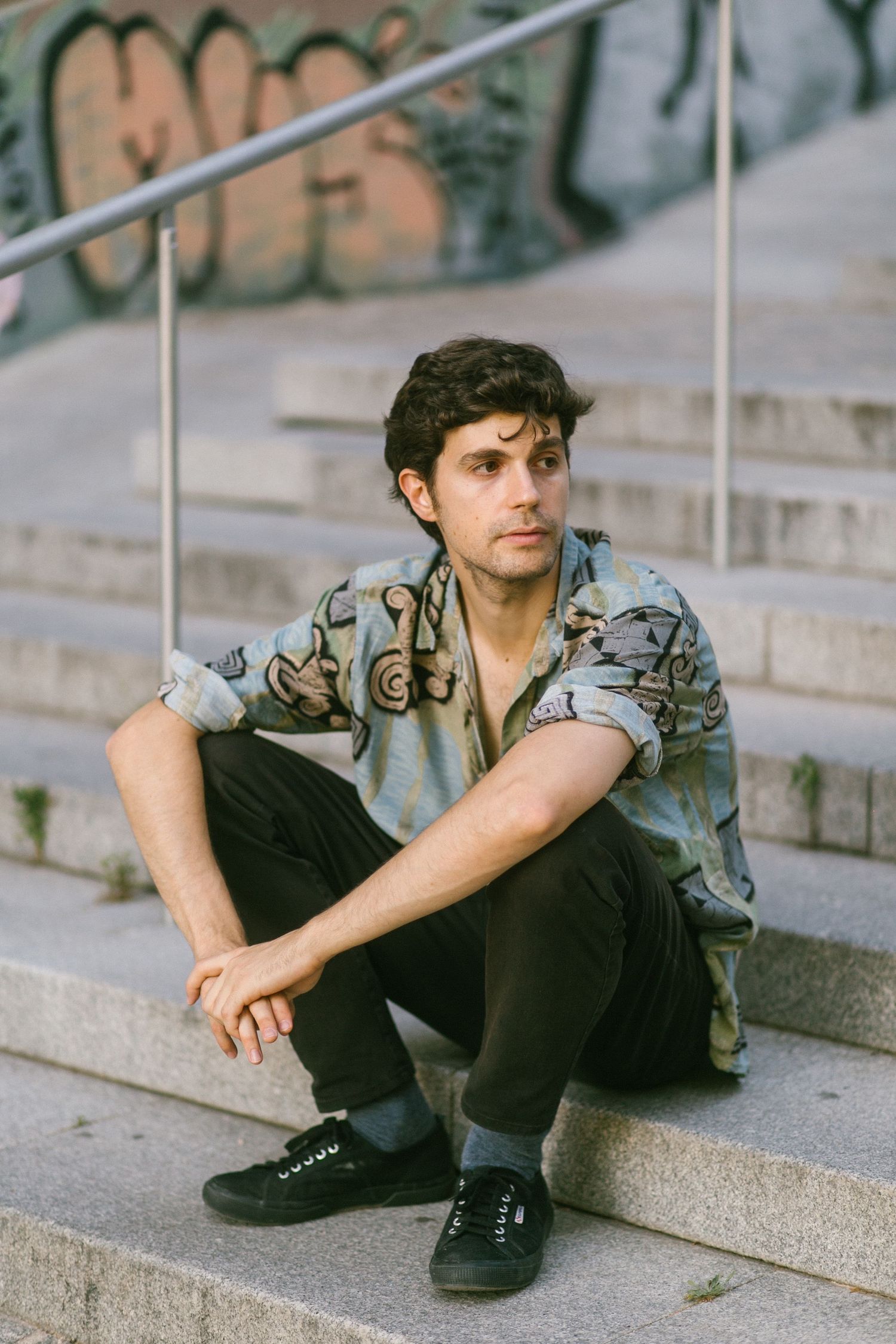A young man in a patterned shirt sits relaxed on stone steps with graffiti wall in background.
