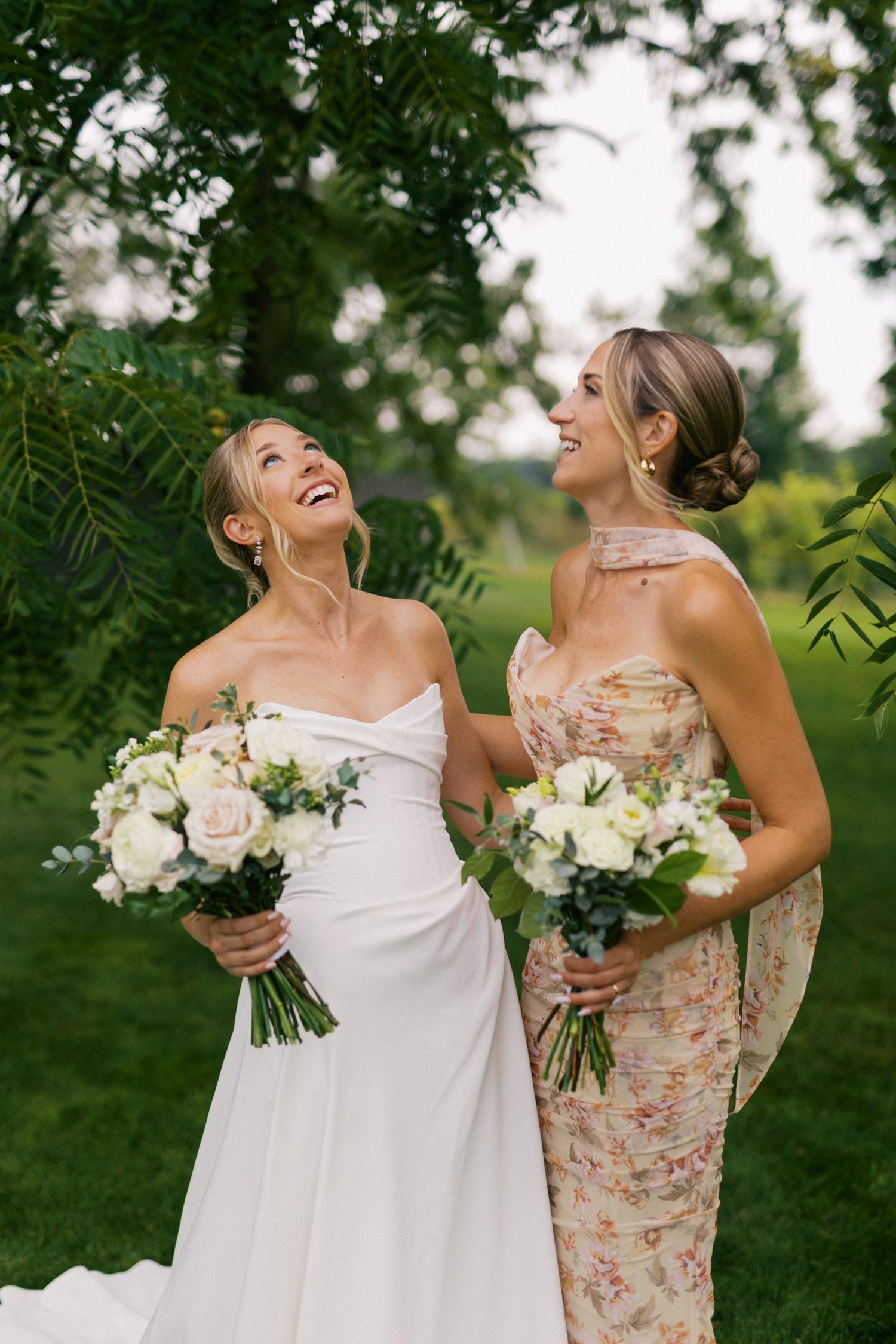 Two wedding party members share joyful laughter while holding white floral bouquets in an outdoor garden setting.