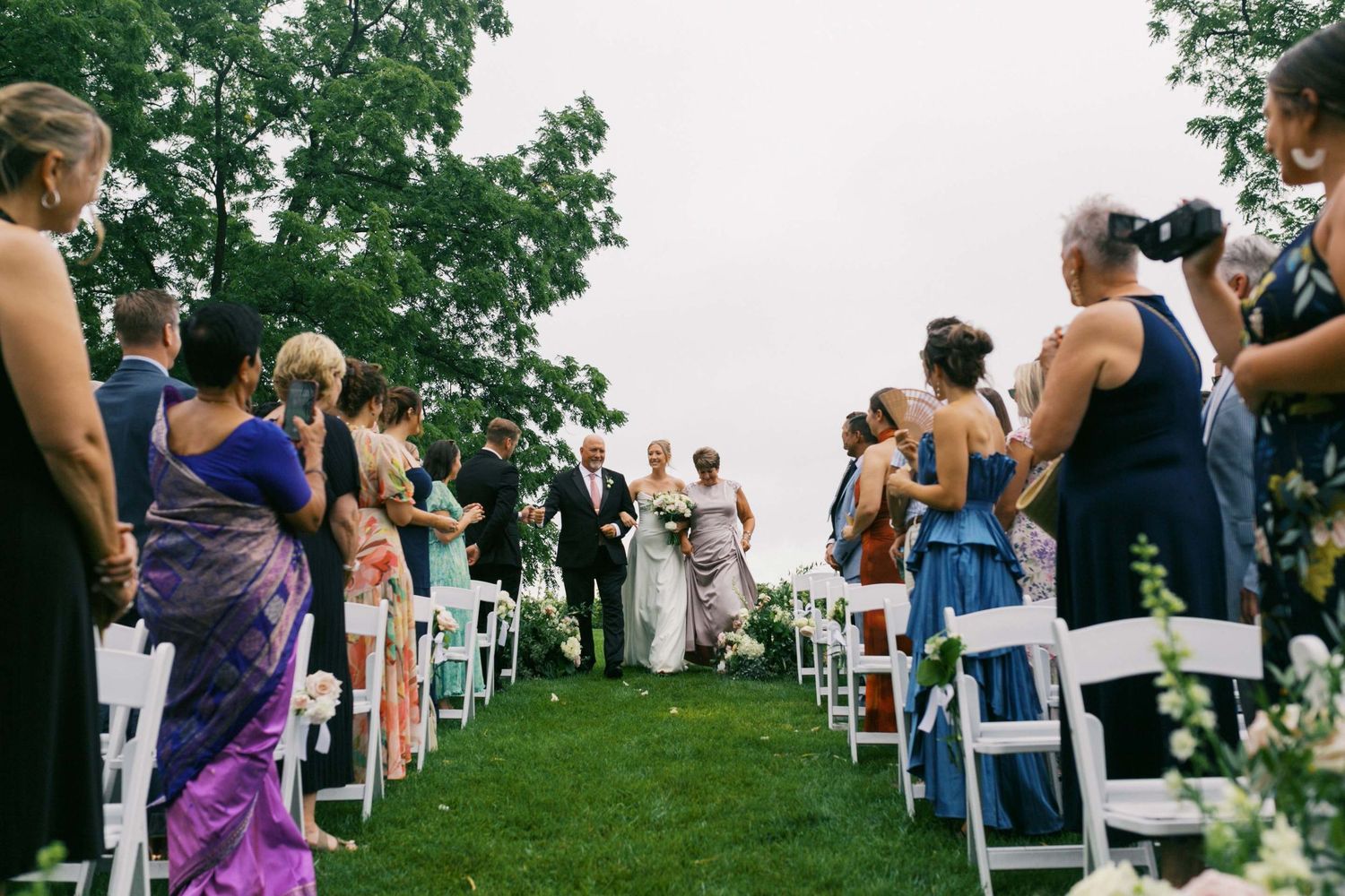 Wedding guests stand and admire a bride and her parents on a grassy hillside during an outdoor ceremony.